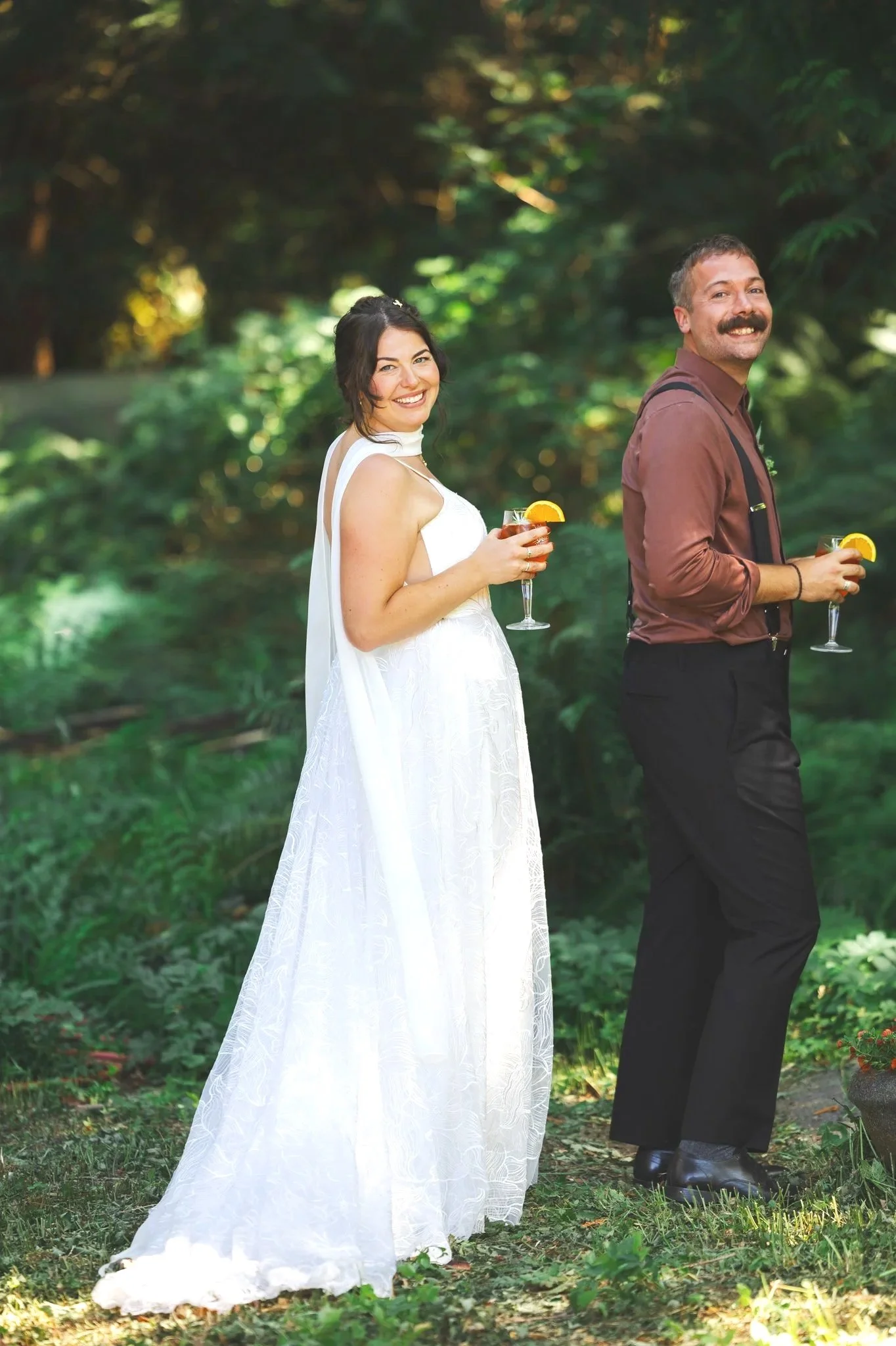A smiling woman in a white dress holding a glass of wine, standing outdoors with a man dressed in a brown shirt and black pants, both holding drinks with lemon wedges, in a lush green forest setting.