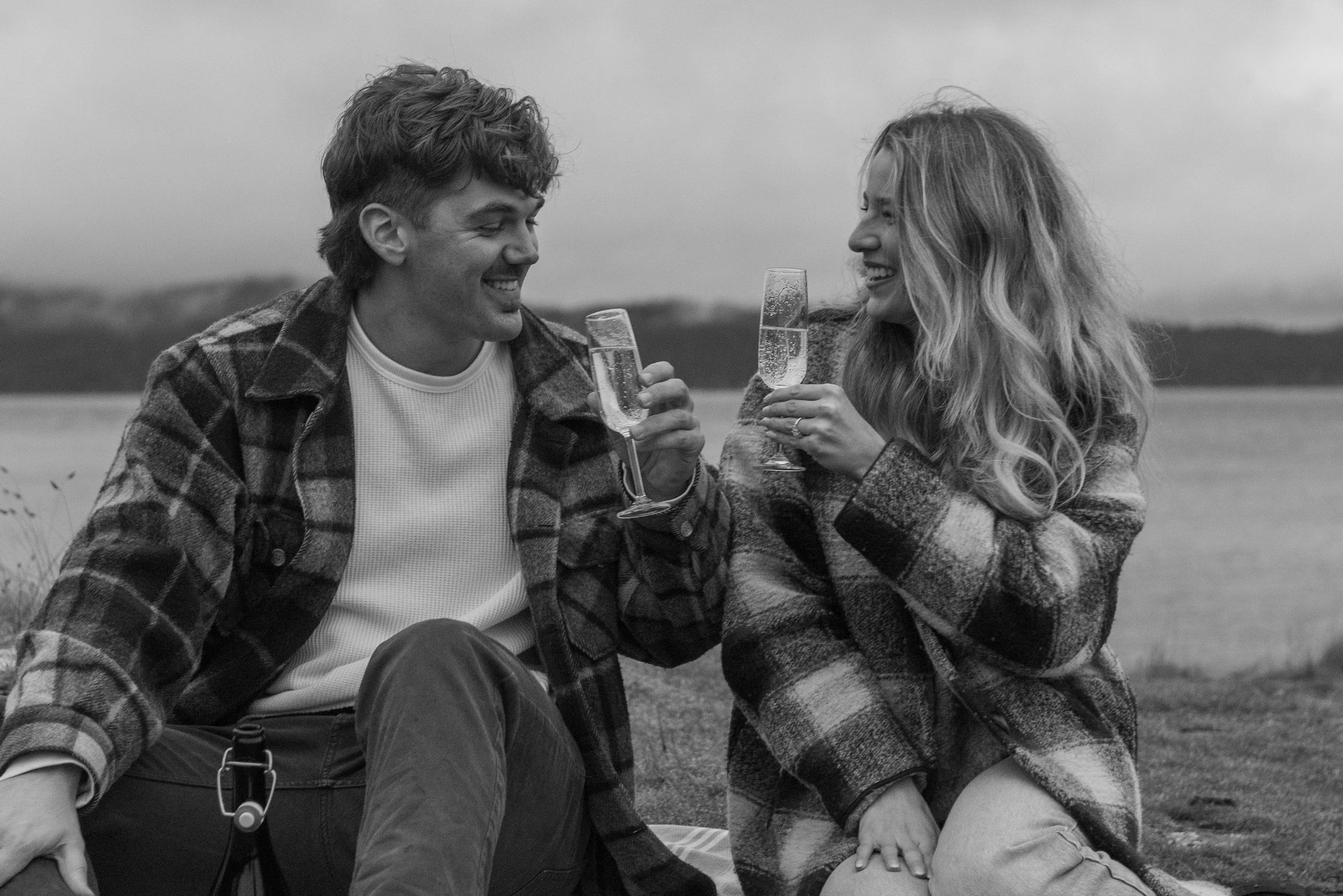 A black and white photo of a man and woman sitting outdoors near a body of water, smiling and clinking glasses of champagne.