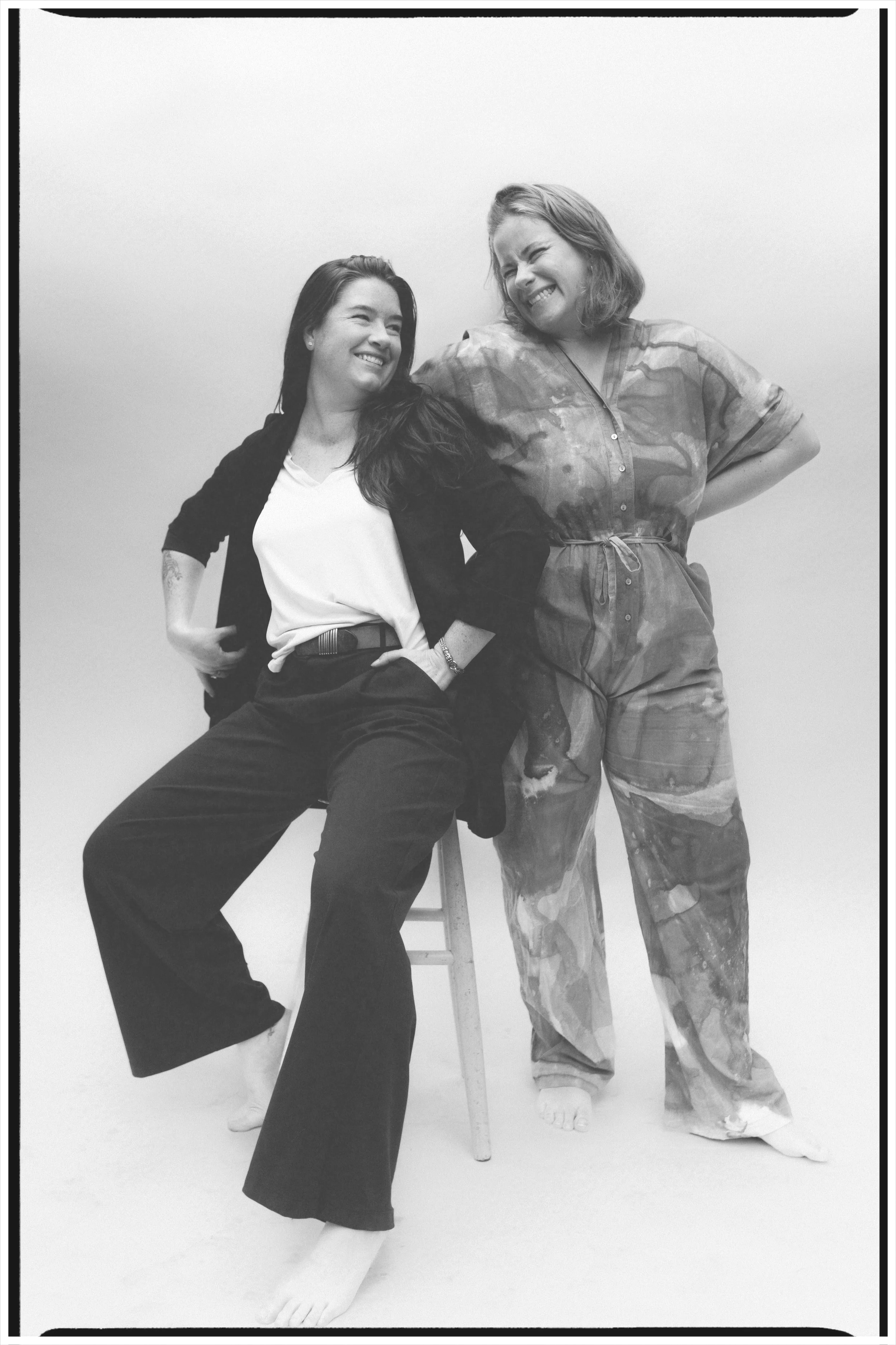Two women smiling and laughing together, one sitting on a stool and the other standing, in a casual setting with a plain background.