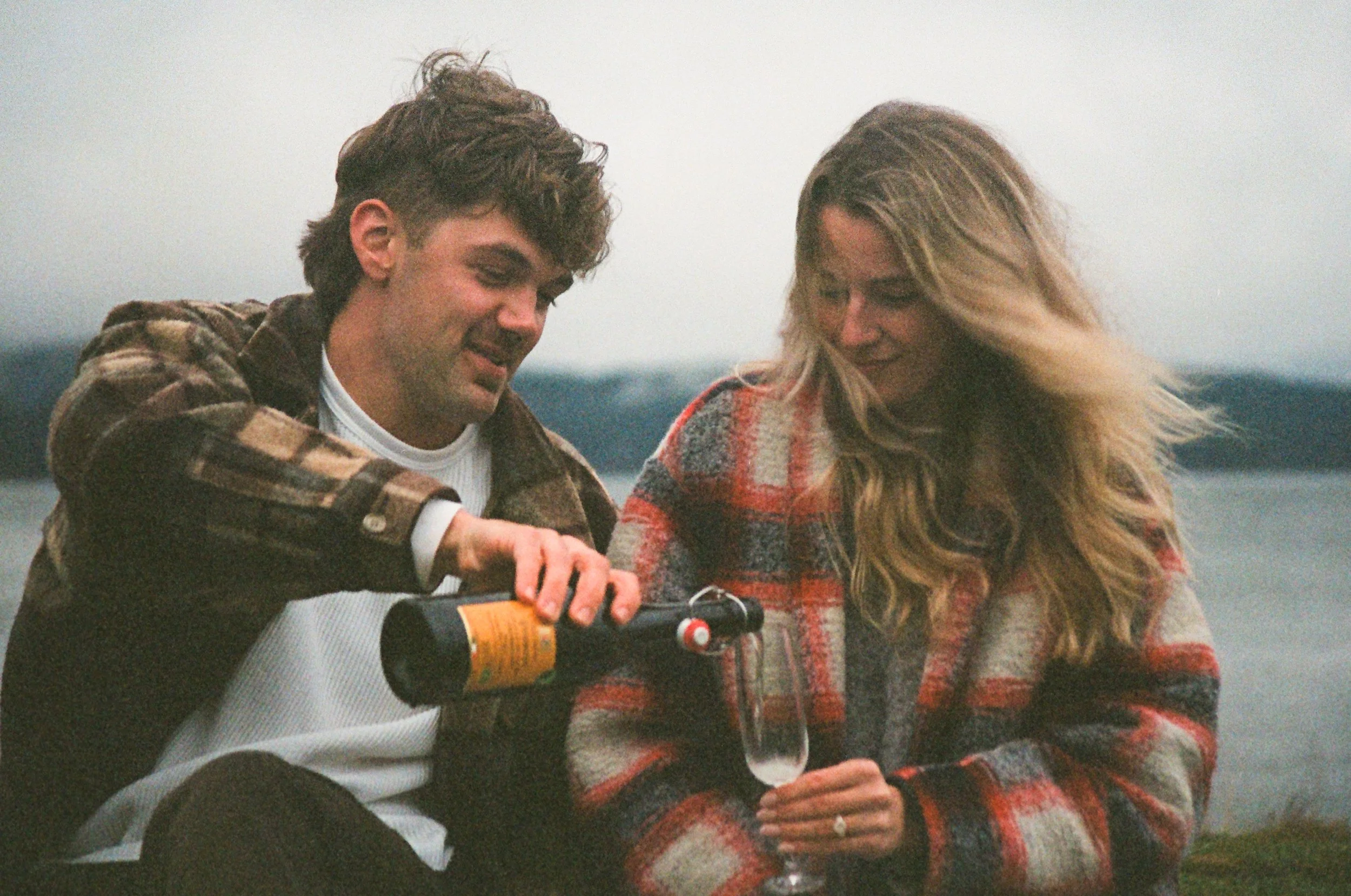 A man and woman sitting outdoors near a body of water, filling a glass of champagne for a toast, both wearing plaid jackets and smiling.