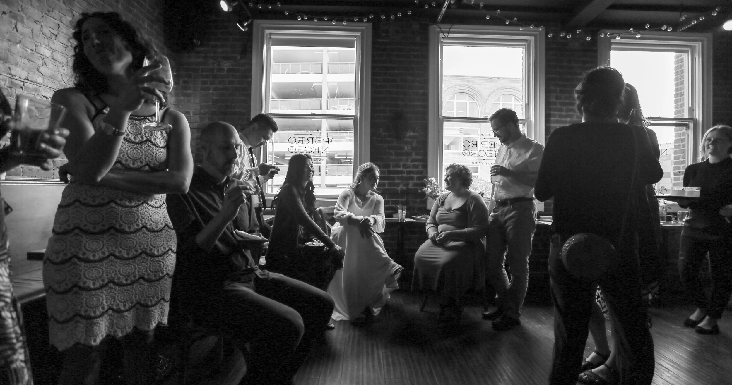 Group of people at a social gathering in a brick-walled restaurant or cafe with large windows, some sitting and some standing, holding drinks and engaging in conversation.