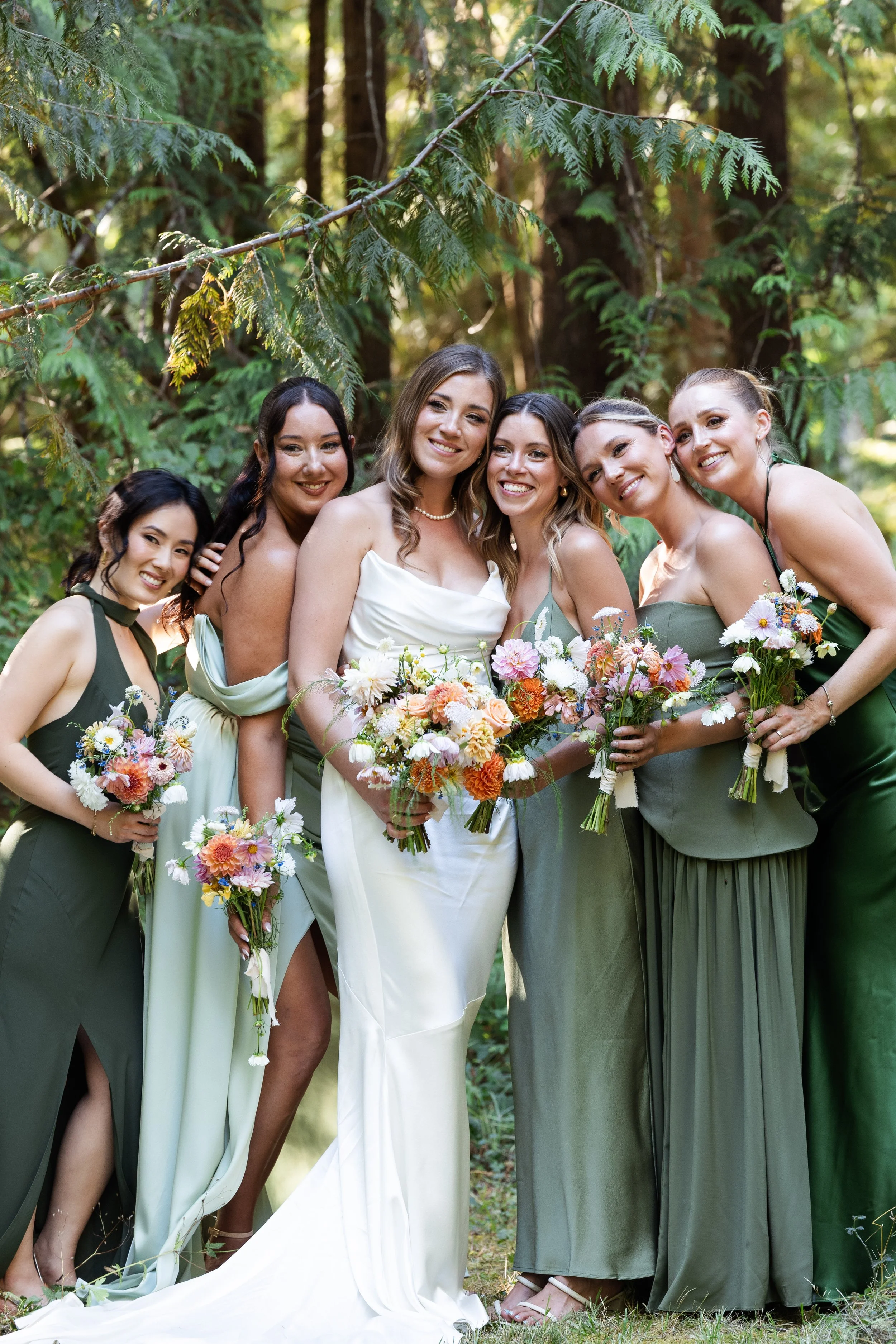 A group of bridesmaids and a bride posing outdoors in a forest, holding bouquets of flowers, smiling for a photo.