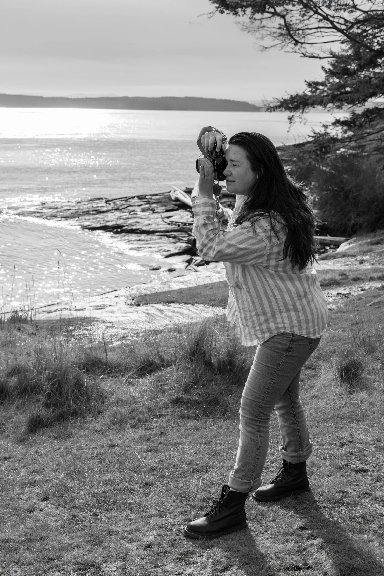 Woman using a camera by a lake with trees in the background, black and white photo.
