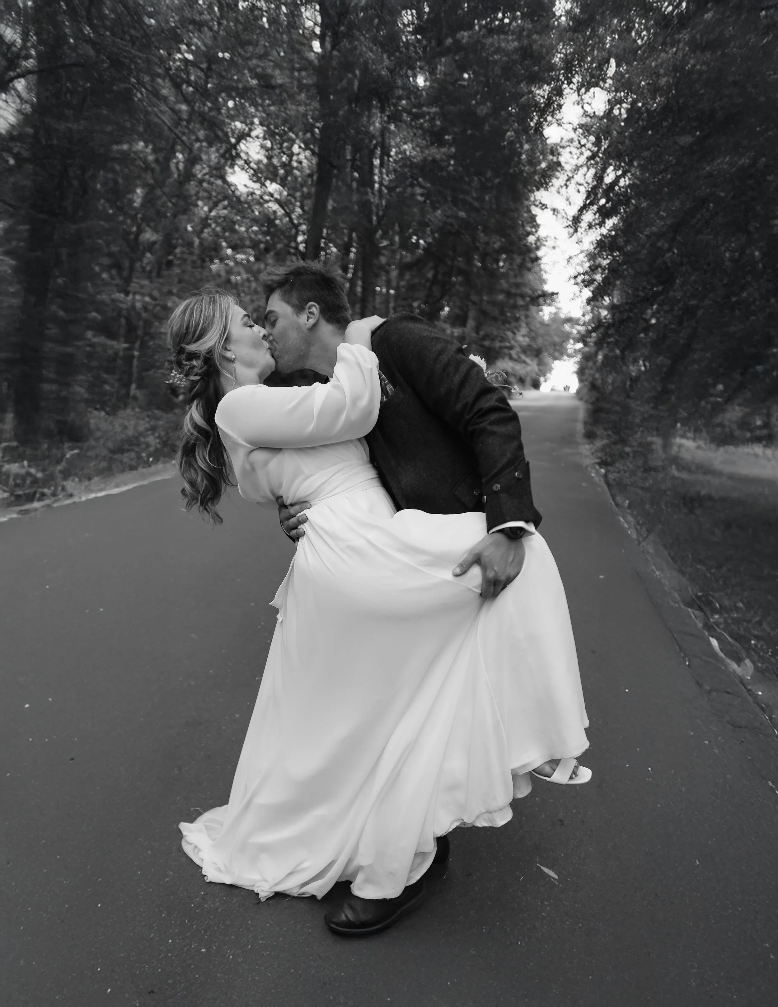 A black and white photo of a couple in wedding attire sharing a kiss on an outdoor road surrounded by trees, with the man holding the woman in a dip.