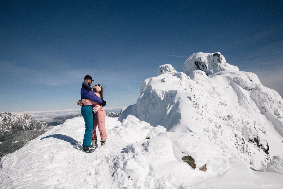 A couple in winter clothing embracing on a snow-covered mountain peak with large snow-covered rocks and a distant mountain range under a clear blue sky.