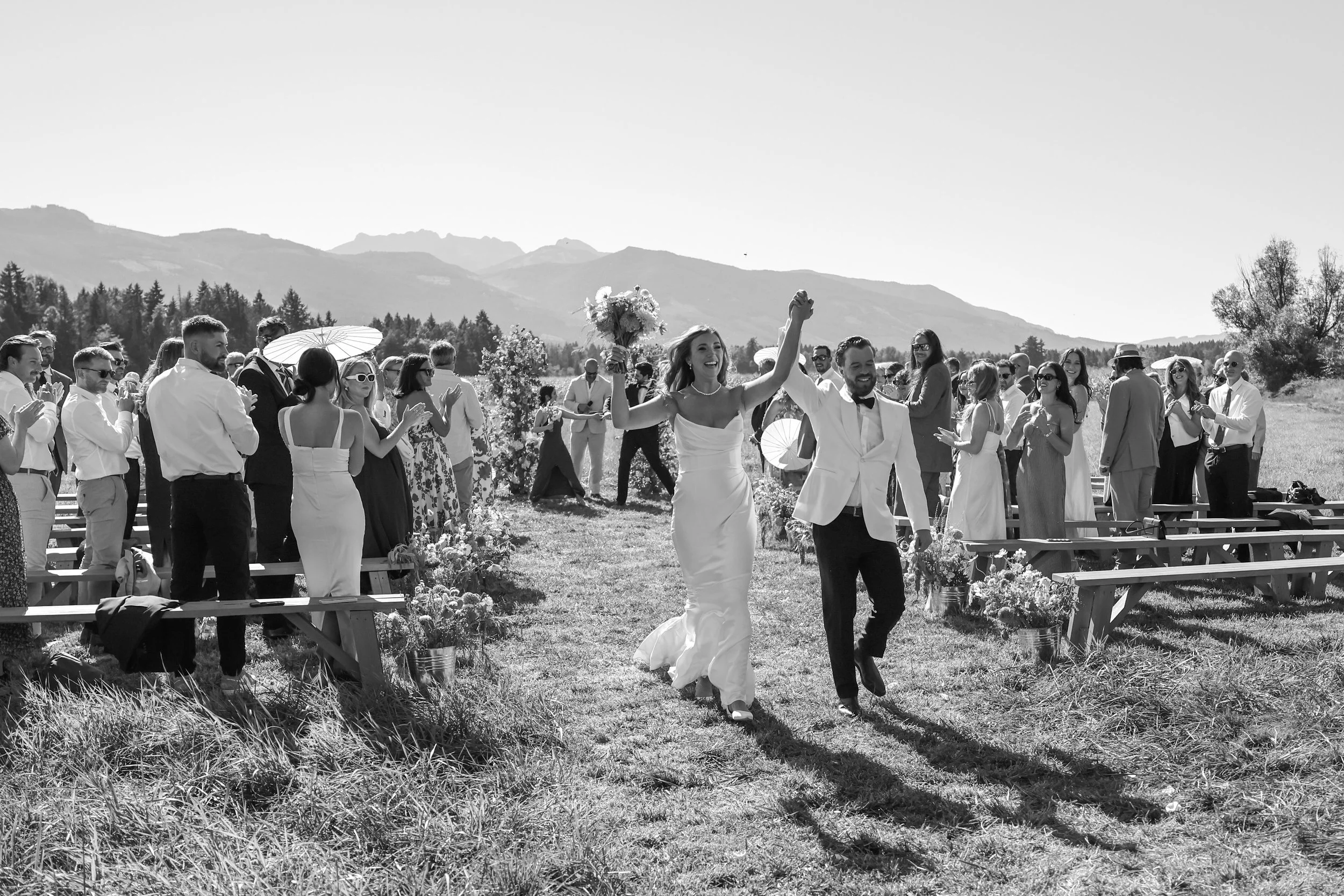 A newlywed couple holding hands and celebrating as they walk down the aisle after a wedding ceremony outdoors, surrounded by friends and family.