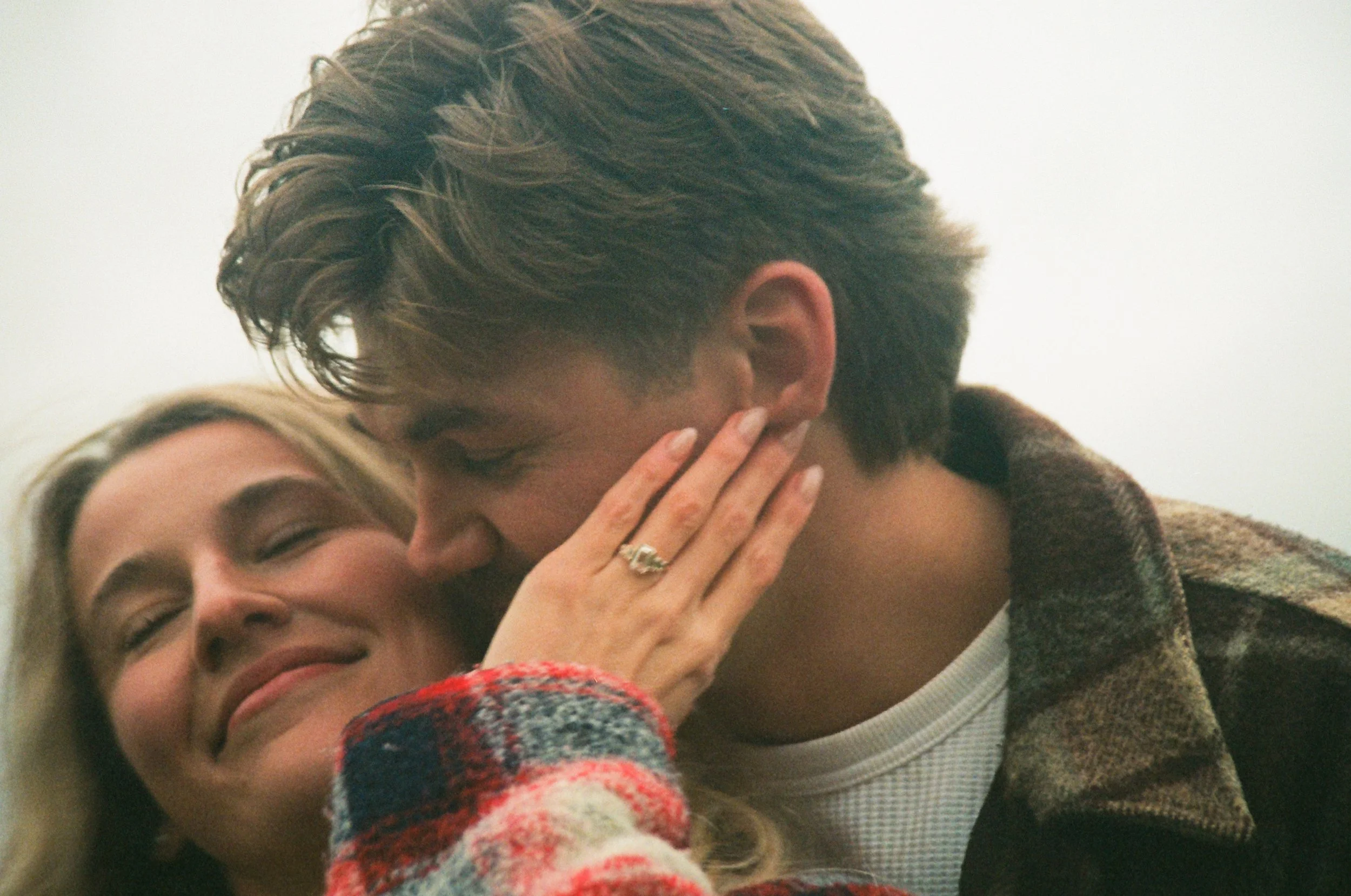 A woman with blonde hair smiling as a man kisses her cheek, her hand touching his face, wearing a ring.