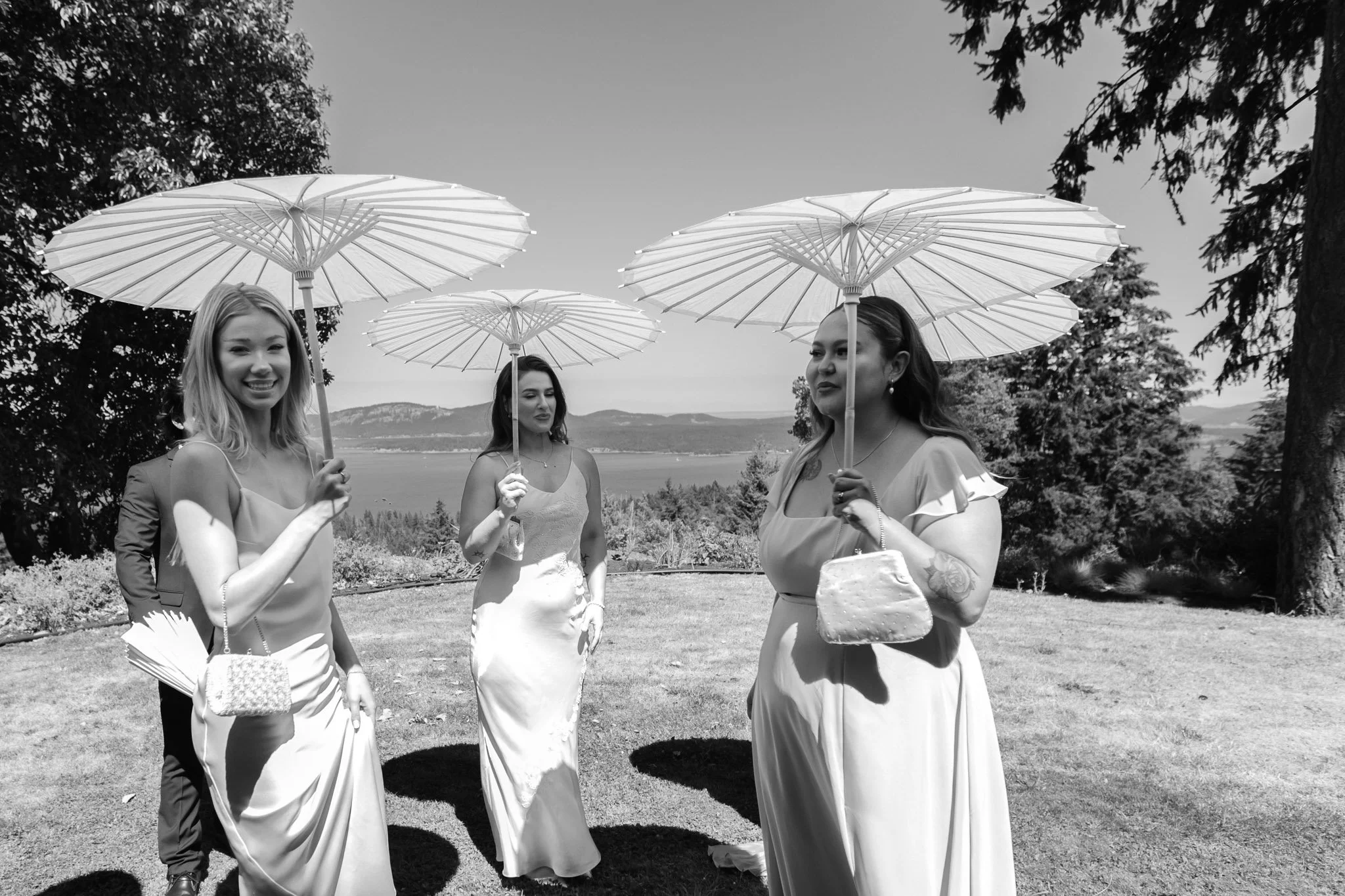 Four women wearing dresses and holding parasols standing outdoors with trees and a body of water in the background. Coast and Kin Wedding Photographer BC Wedding Photographer, Salt Spring Island Wedding Photographer, Vancouver Island Wedding