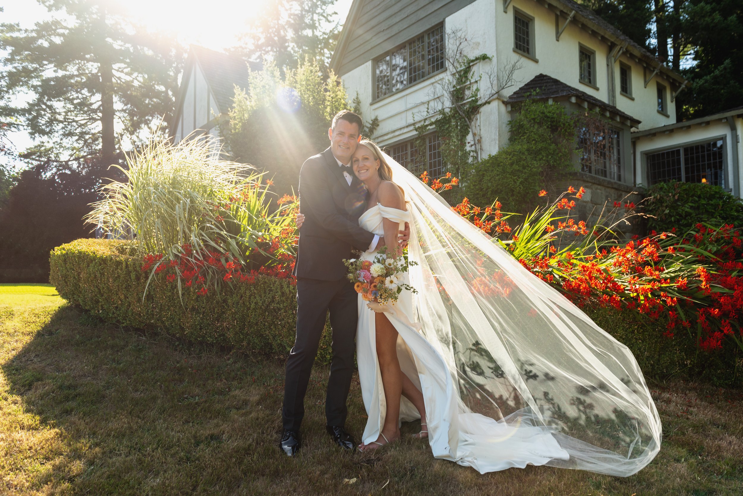 A bride and groom embracing outdoors on their wedding day, with sunlight shining behind them and a historic house in the background, surrounded by colorful flowers and lush greenery.