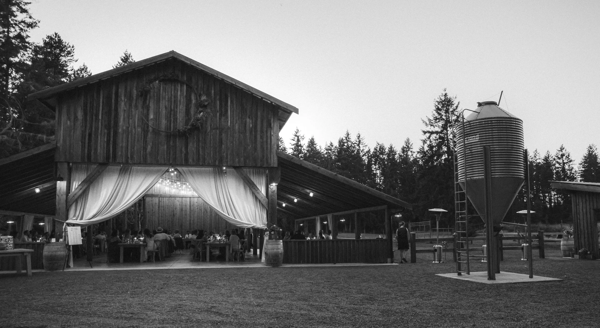 Outdoor barn-style wedding reception with guests seated at tables inside a decorated open-air structure, with string lights and draped curtains, set against a backdrop of trees at dusk.