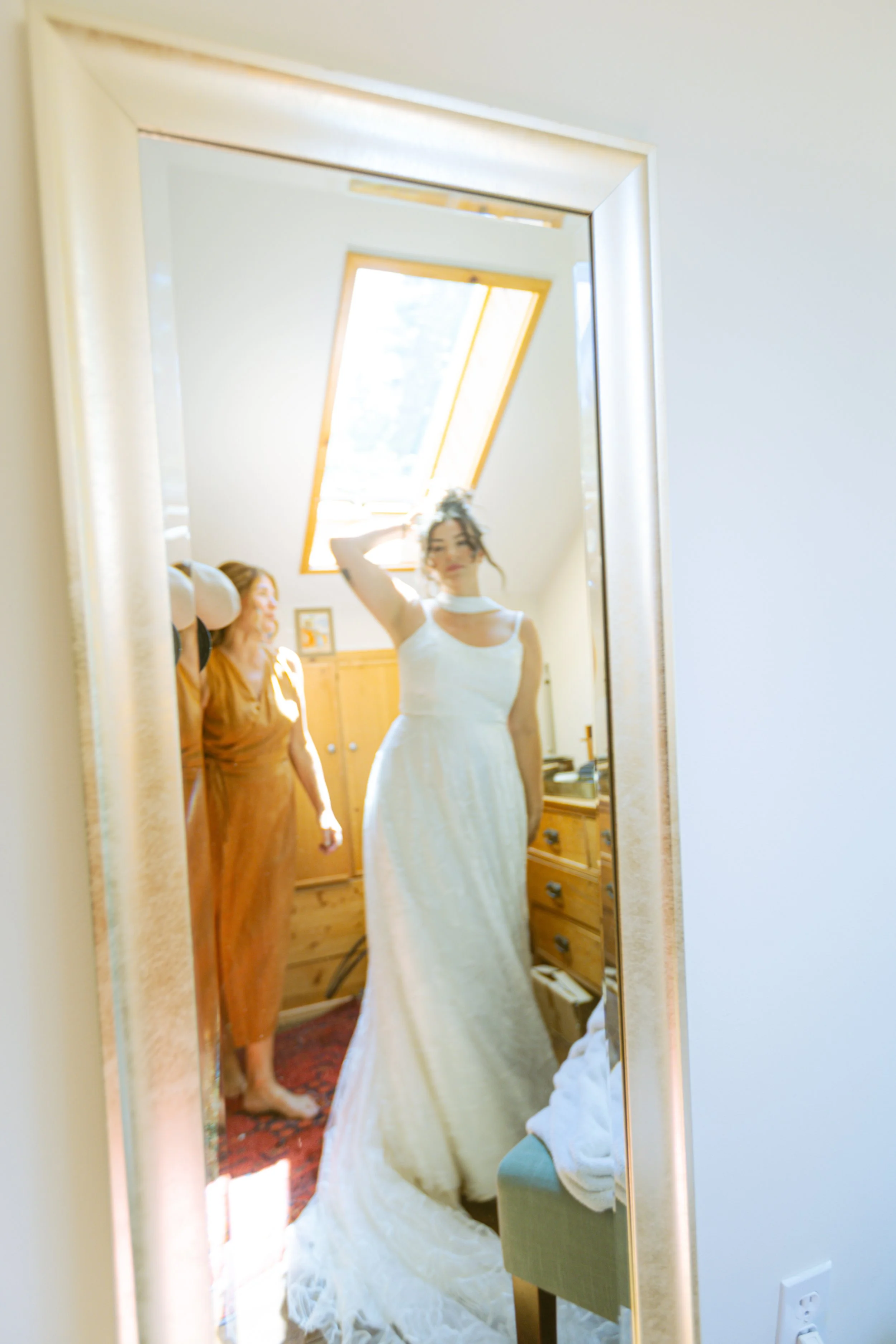 A woman in a white wedding dress taking a selfie in a mirror, with another woman in a brown dress standing nearby in a bright room with a skylight.