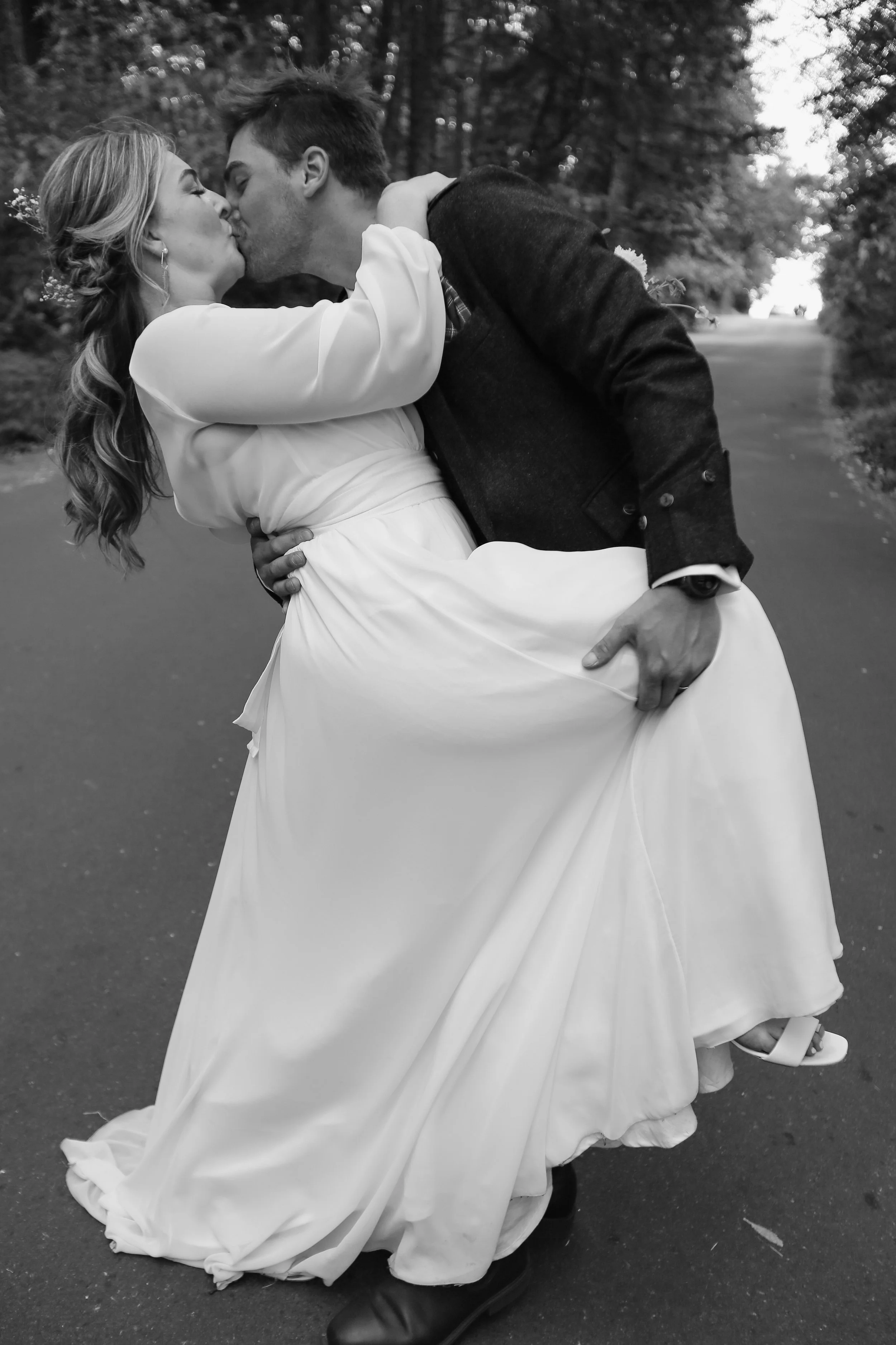 A black-and-white photo of a man and woman kissing outdoors, with the man lifting the woman in his arms. The woman is wearing a long white dress, and the man is dressed in a dark jacket and pants. They are standing on a paved path surrounded by trees