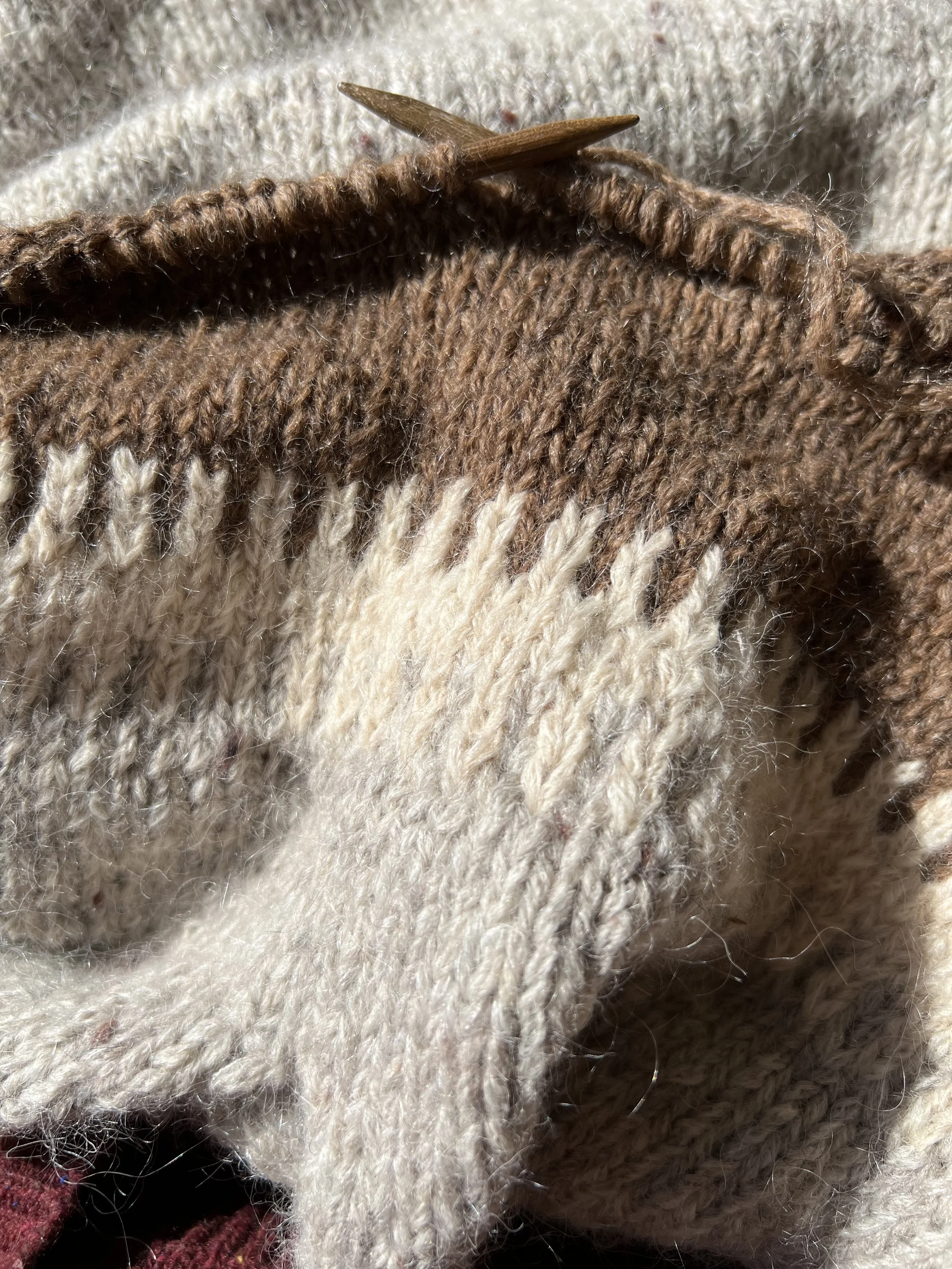 Close-up of a knitted sweater with brown, cream, and beige yarn, and a wooden knitting needle inserted into the fabric.