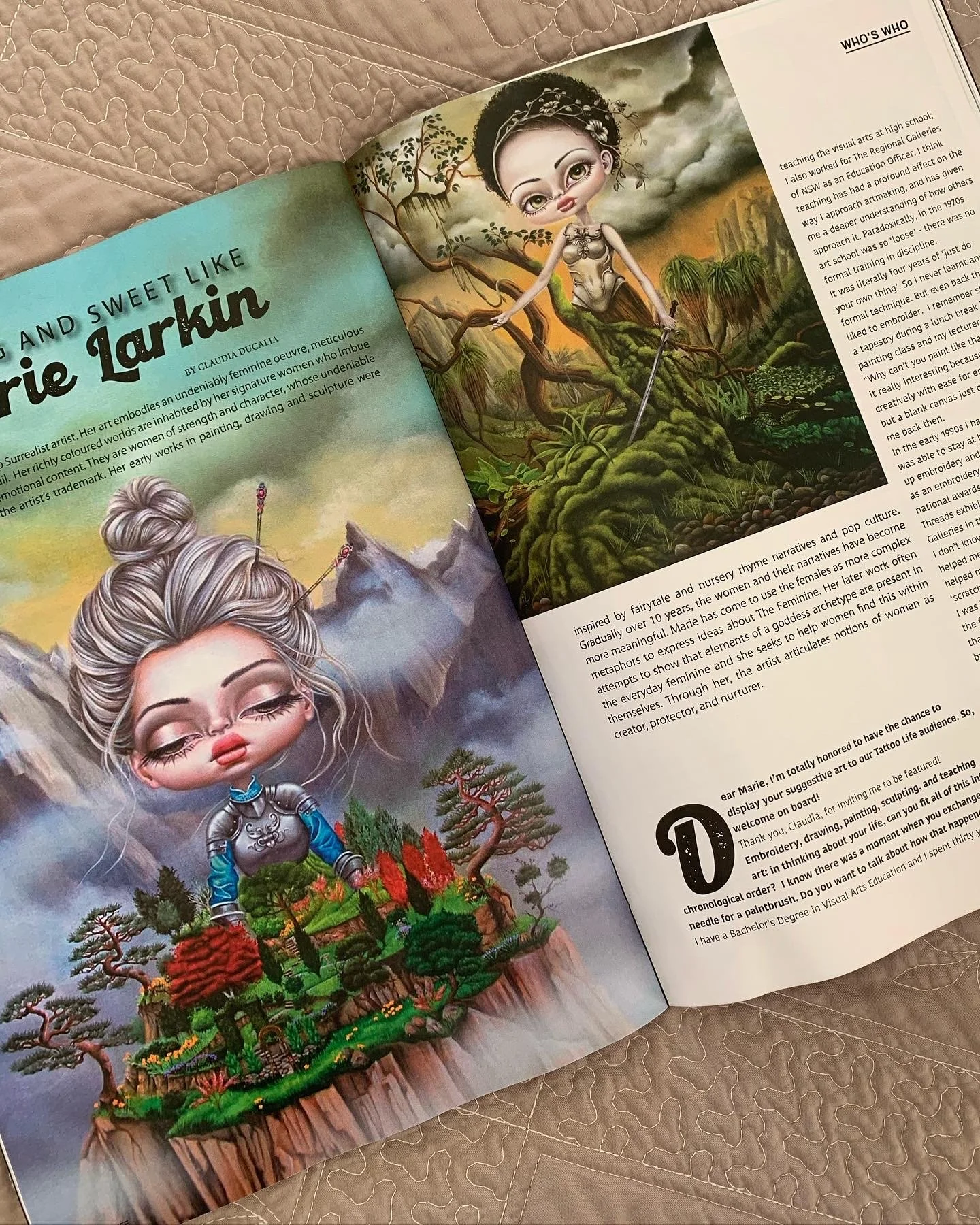 An open magazine on a bed features illustrated artworks of Marie Larkin, a female character in fantasy style. The left page shows a cartoon girl with long, gray hair styled in a bun, dressed in armor, standing in a lush landscape with trees and mountains. The right page depicts a fairy-tale inspired landscape with a whimsical female figure with large eyes, dark hair, and an elaborate outfit, set against a stormy sky and green hills.