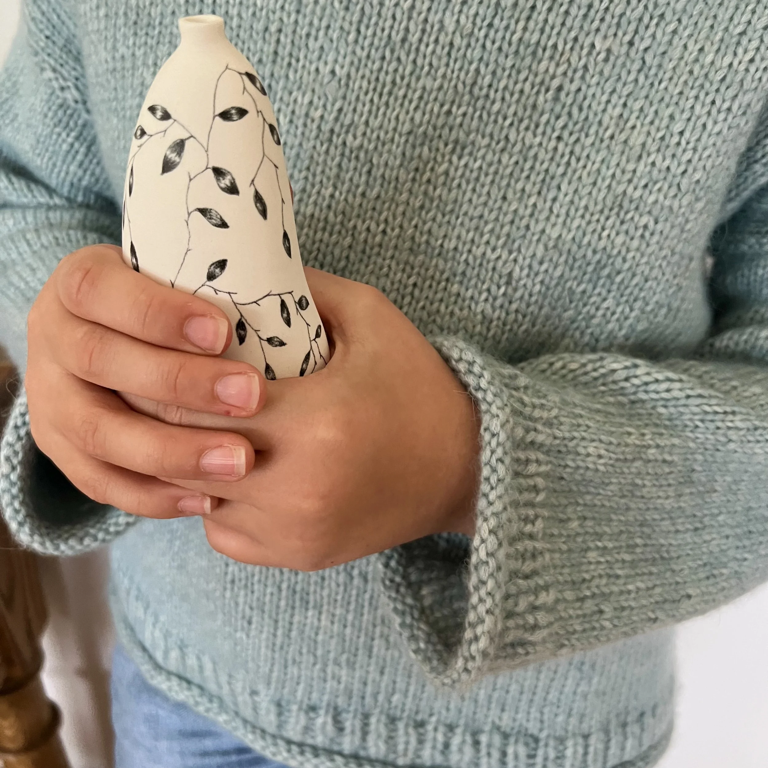 Close up young girl hands holding a small ceramic black and white vase with leaves on it wearing an aqua blue hand knit sweater.