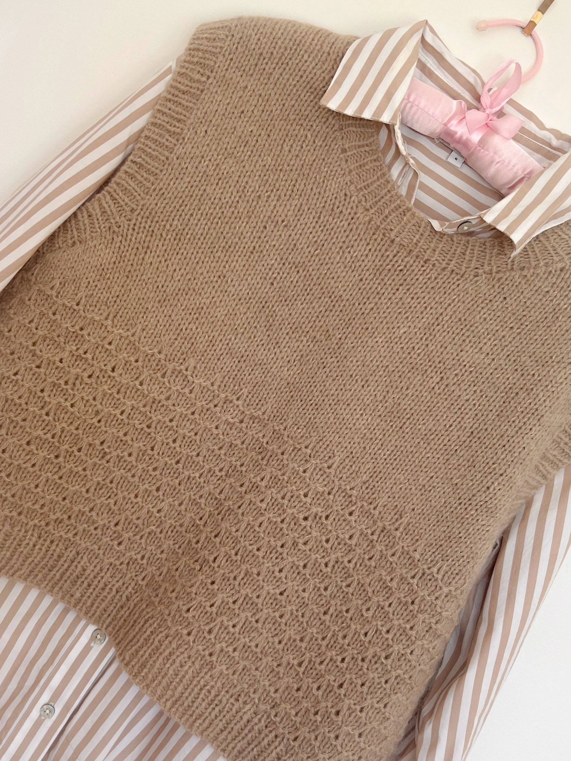 Mid brown hand knit vest with decorative stitches hanging on a pink coat hanger.