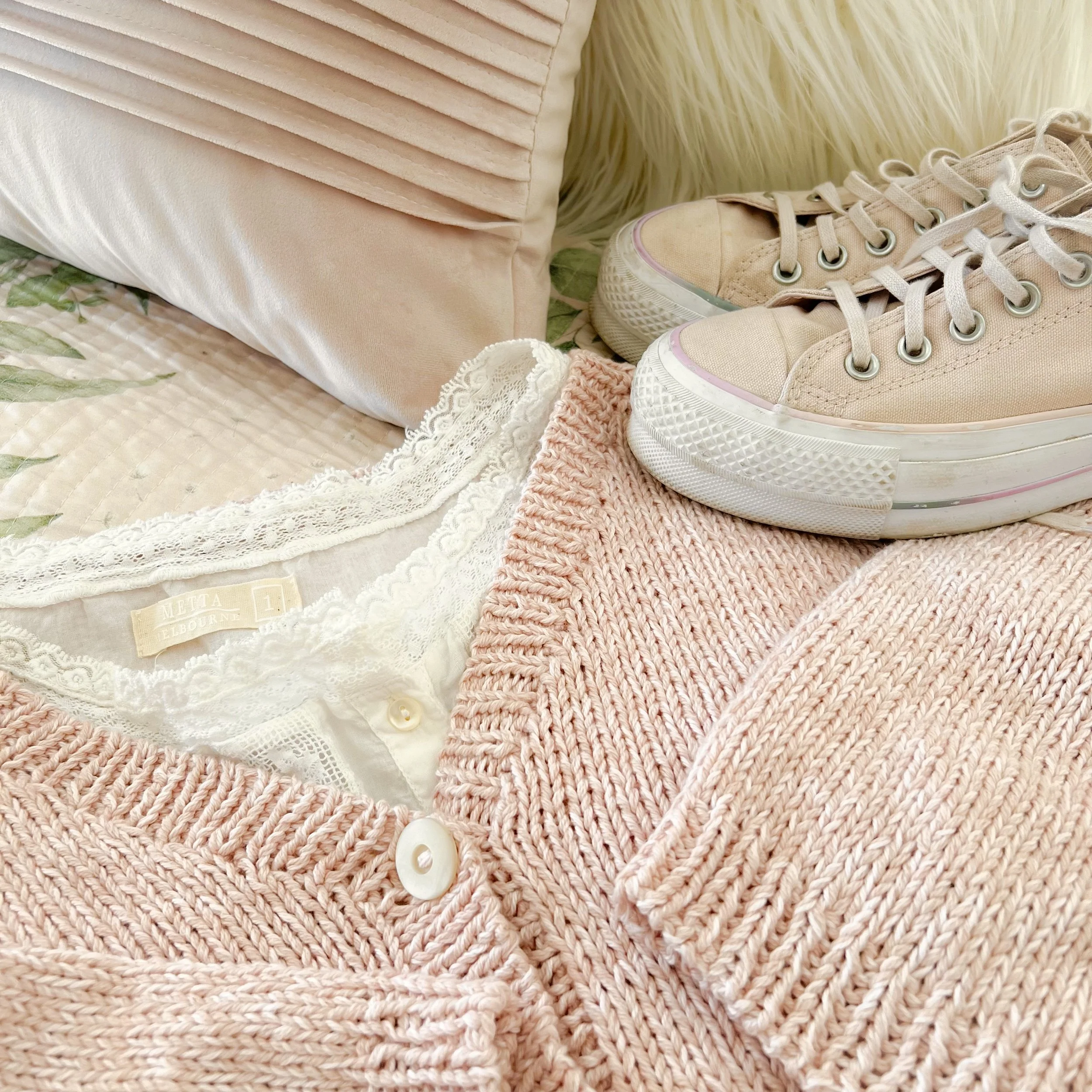 Handknit pale pink cotton cardigan with a white shirt with lace edging styled next to lace pink sneakers and a pale pink cushion.
