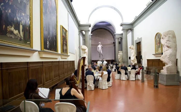 Elegant banquet in a museum hall with a statue and large paintings, featuring a harpist and guests seated at round tables.