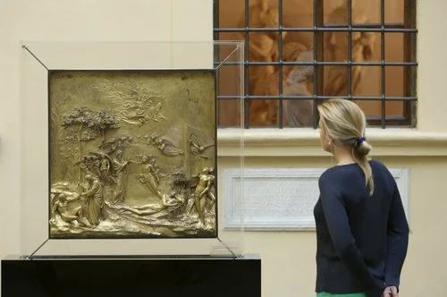 A woman observing a gilded bronze relief art piece displayed in a museum case, with a barred window in the background.