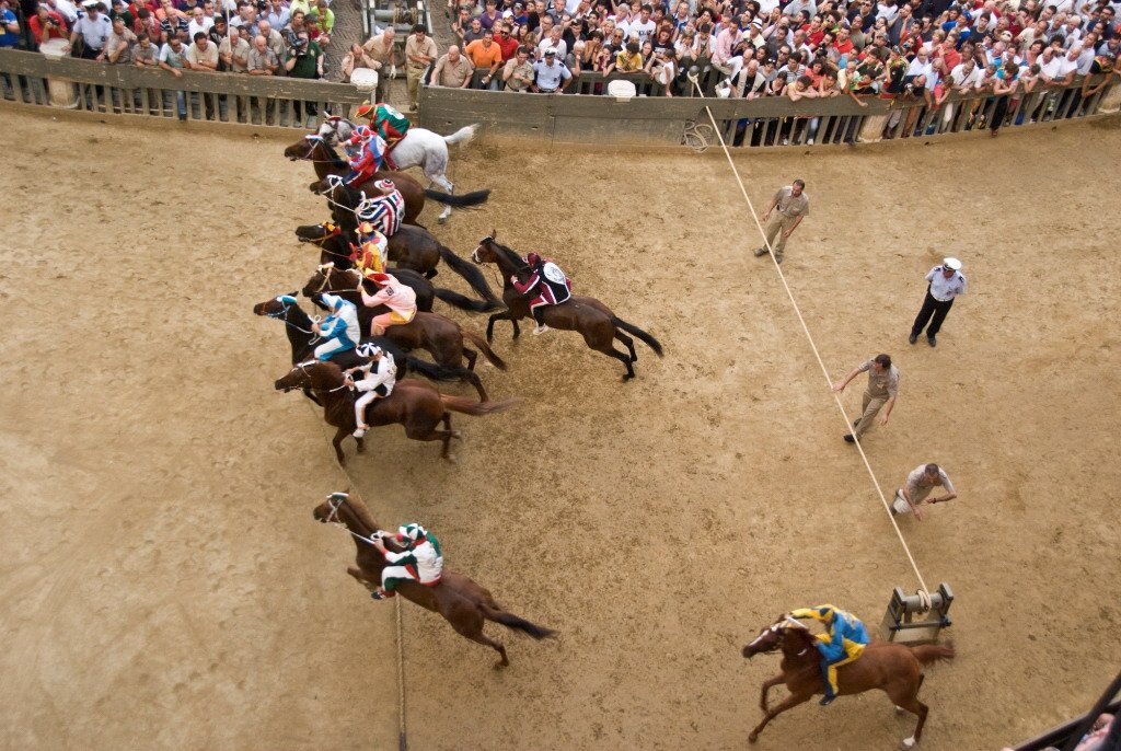 Horse race starting, jockeys wearing colorful uniforms, crowded spectators behind a barrier, seen from above.