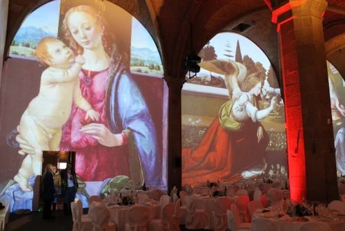 Interior of a hall with large art projections on walls featuring Renaissance paintings, including Madonna and Child and an angel, with tables and chairs set for an event.