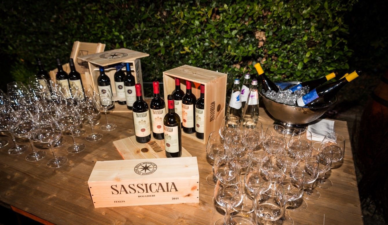 Wine bottles, wooden crates, wine glasses, and a metal ice bucket with bottled drinks on a wooden table outdoors.