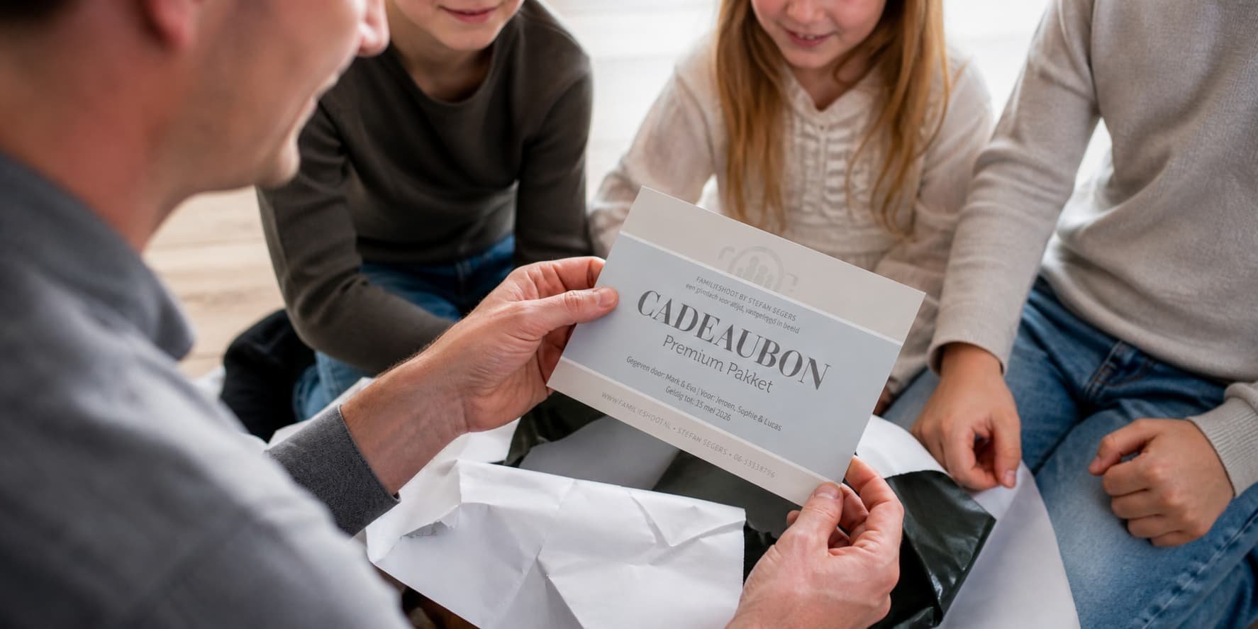 Een familie zit samen en kijkt naar een cadeaubon die door een volwassene wordt vastgehouden. De cadeaubon is van het merk Cadeaubon en is voor een Premium Pakket.