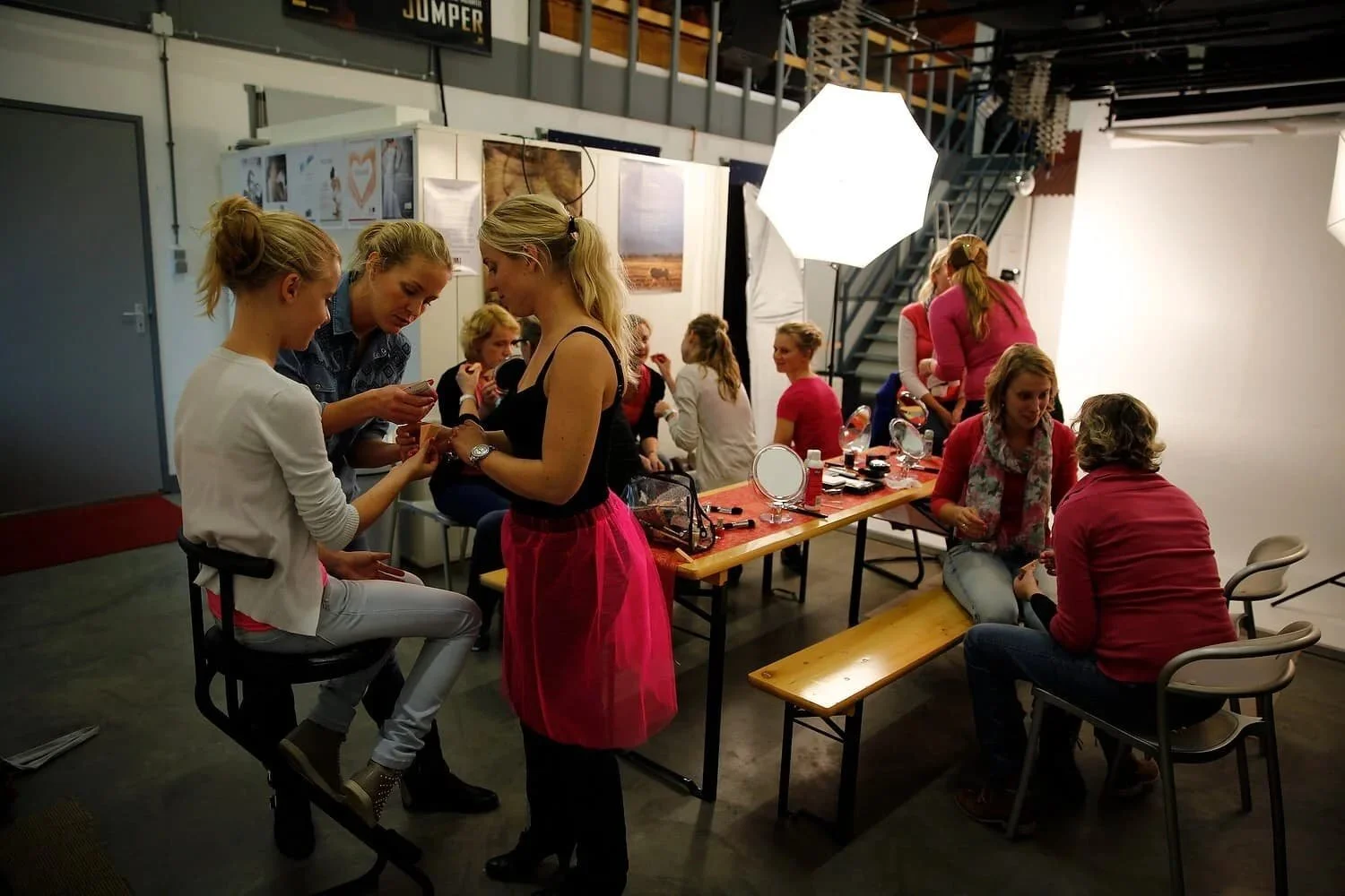 Vrouw die make-up aanbrengt bij een jonge vrouw tijdens een beauty evenement met make-upspiegel, groep vrouwen in de achtergrond, professionele belichting