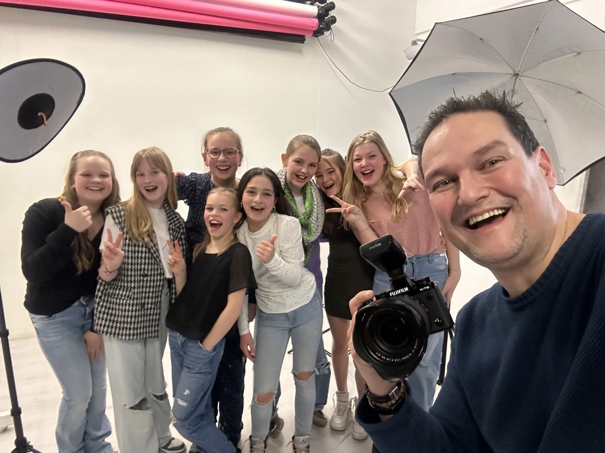 Een groep jonge meisjes poseert voor een foto in een studio, met een man die de camera vasthoudt. De meisjes lachen en maken V-vlaggen met hun handen. De man lacht tevreden aan de rechterkant van de foto.
