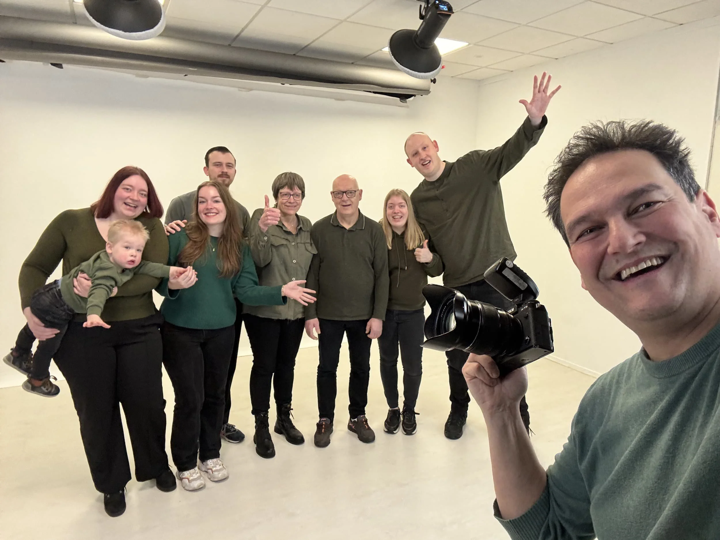 Een groep mensen poseert voor de camera, terwijl één persoon een losse foto maakt. Ze lachen en lijken plezier te hebben in een binnenruimte met een witte muur.