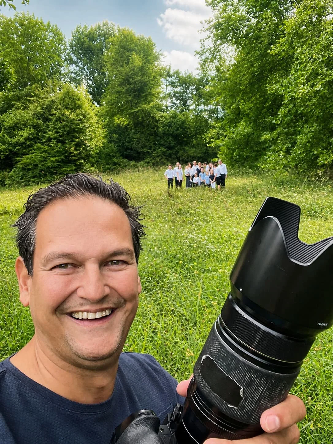 Familiefotograaf Stefan Segers die lachend in de natuur poseert met een grote camera, terwijl op de achtergrond een groep mensen die allemaal in formele kleding staan.