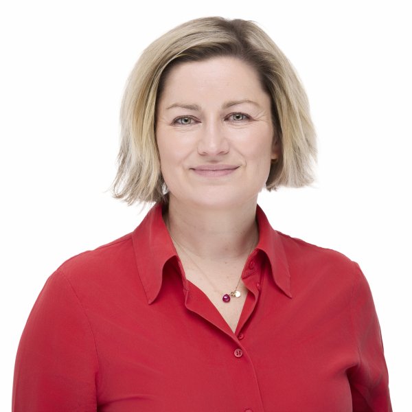 A woman with shoulder-length blonde hair smiling and wearing a red button-up shirt, standing against a plain white background.