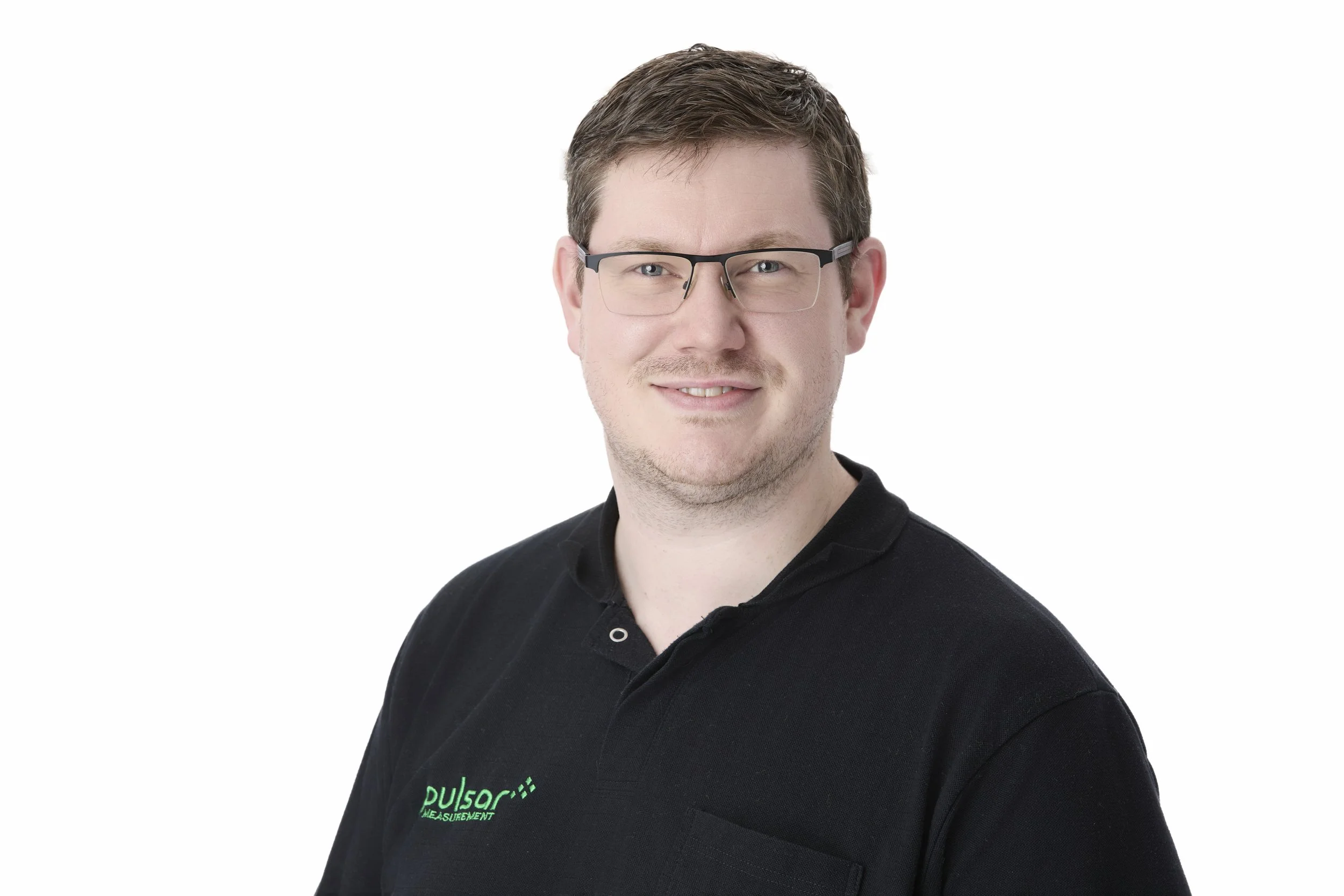 A man with glasses and short brown hair smiling, wearing a black polo shirt with green embroidered text, against a plain white background.