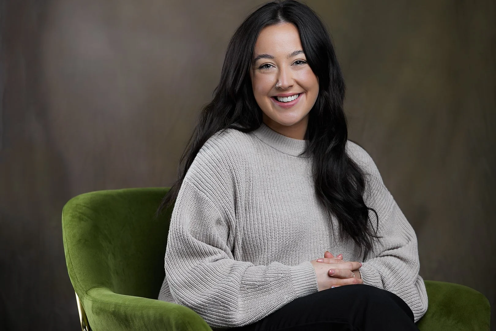 A young woman with dark hair, smiling, sitting on a green chair against a plain background, wearing a light gray sweater.