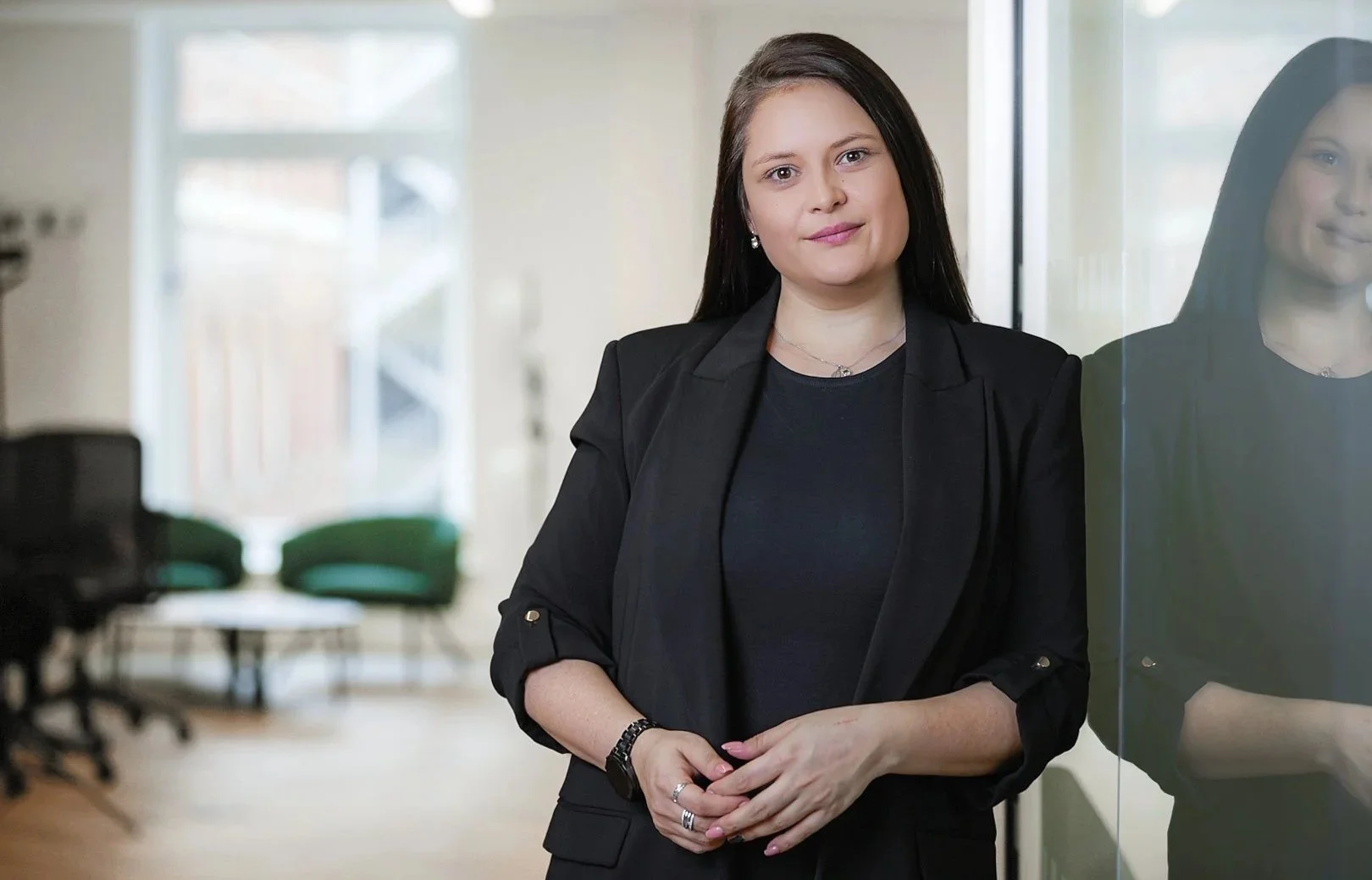 A woman with brown hair, dressed in a black blazer and t-shirt, standing next to a glass wall in a modern office setting.