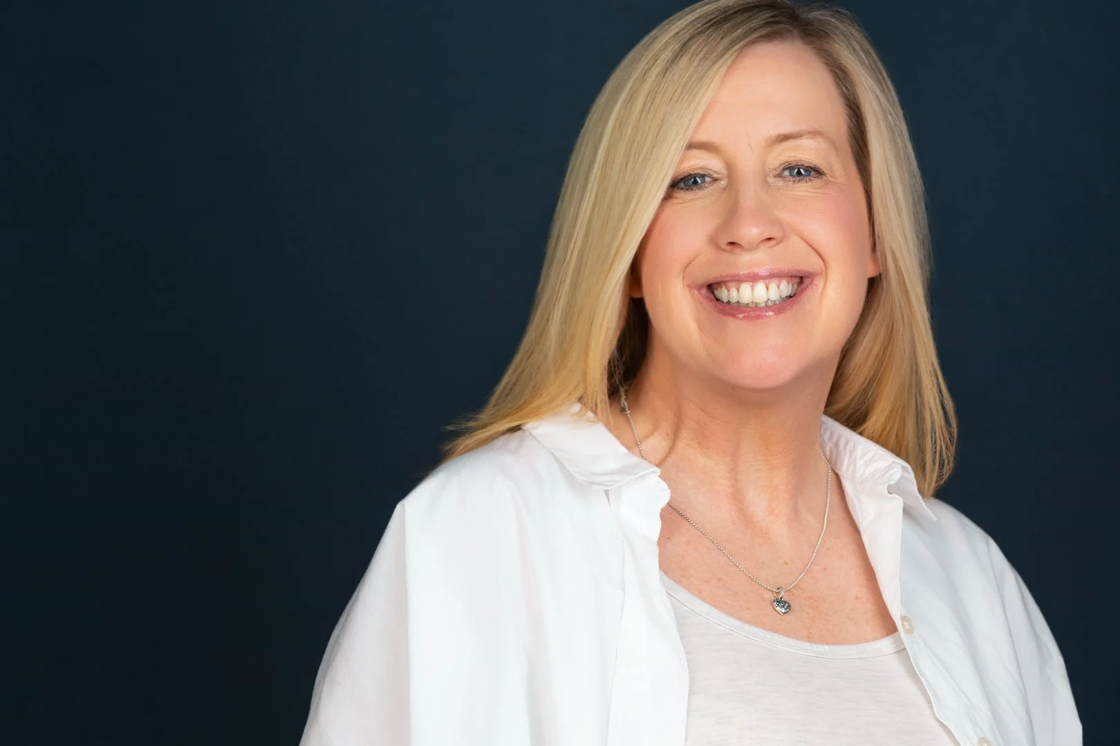 A woman with blonde hair smiling, wearing a white top and necklace, standing against a dark background.