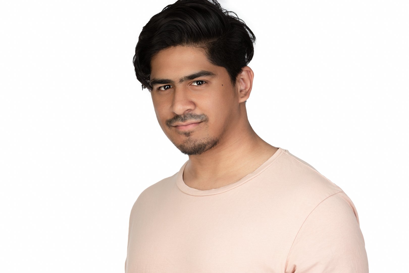 A young man with dark hair and a light beard, wearing a light pink t-shirt, smiling slightly at the camera against a plain white background.