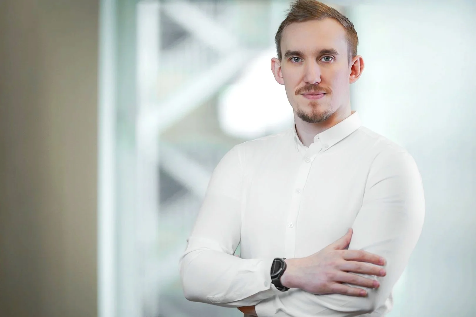 A confident man with short hair and a beard, wearing a white dress shirt and a black watch, standing with arms crossed in a modern office environment.