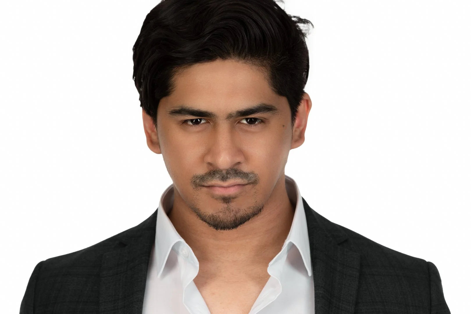 A confident man with dark hair and a well-groomed beard wearing a black suit and white shirt, looking directly at the camera against a plain white background.