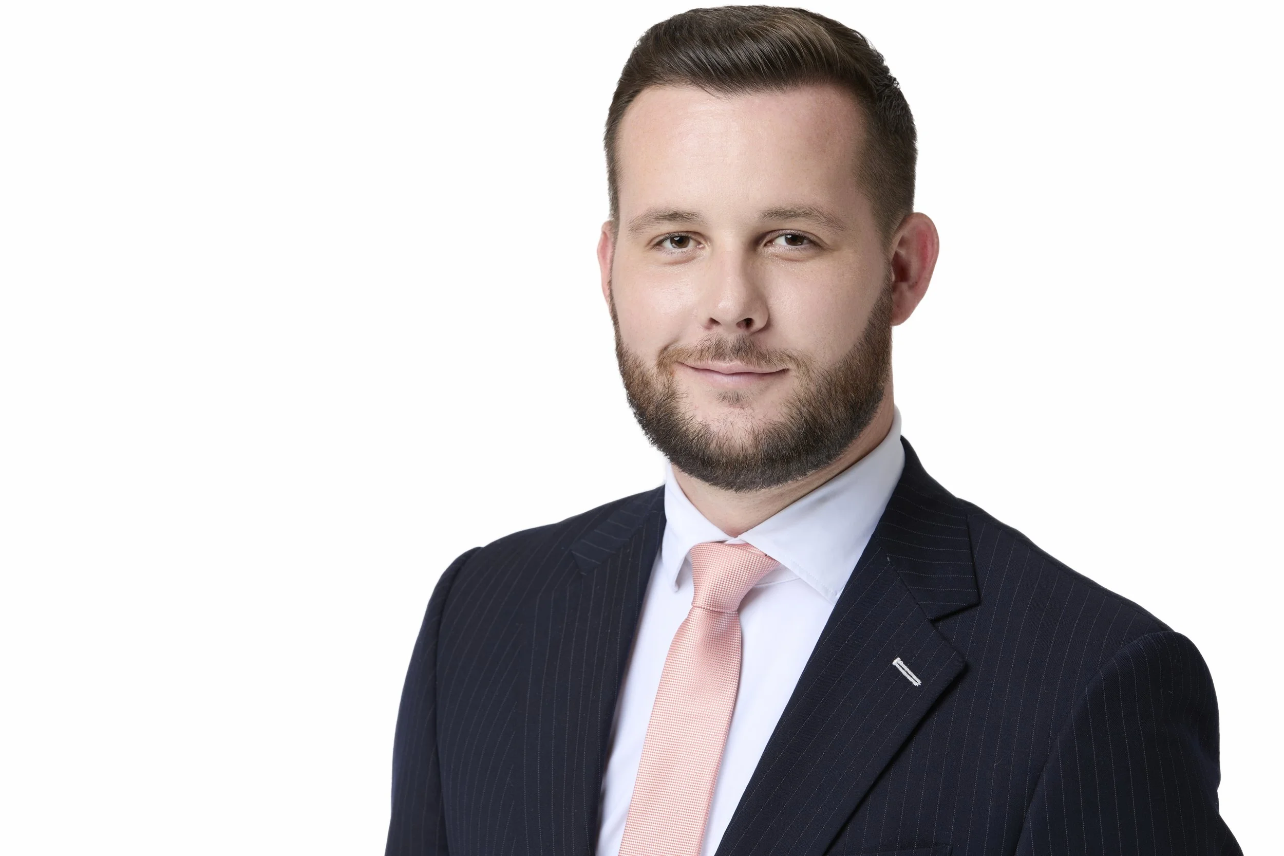 Professional portrait of a man with short brown hair and a beard, wearing a navy pinstripe suit, white shirt, and light pink tie, smiling confidently against a plain white background.