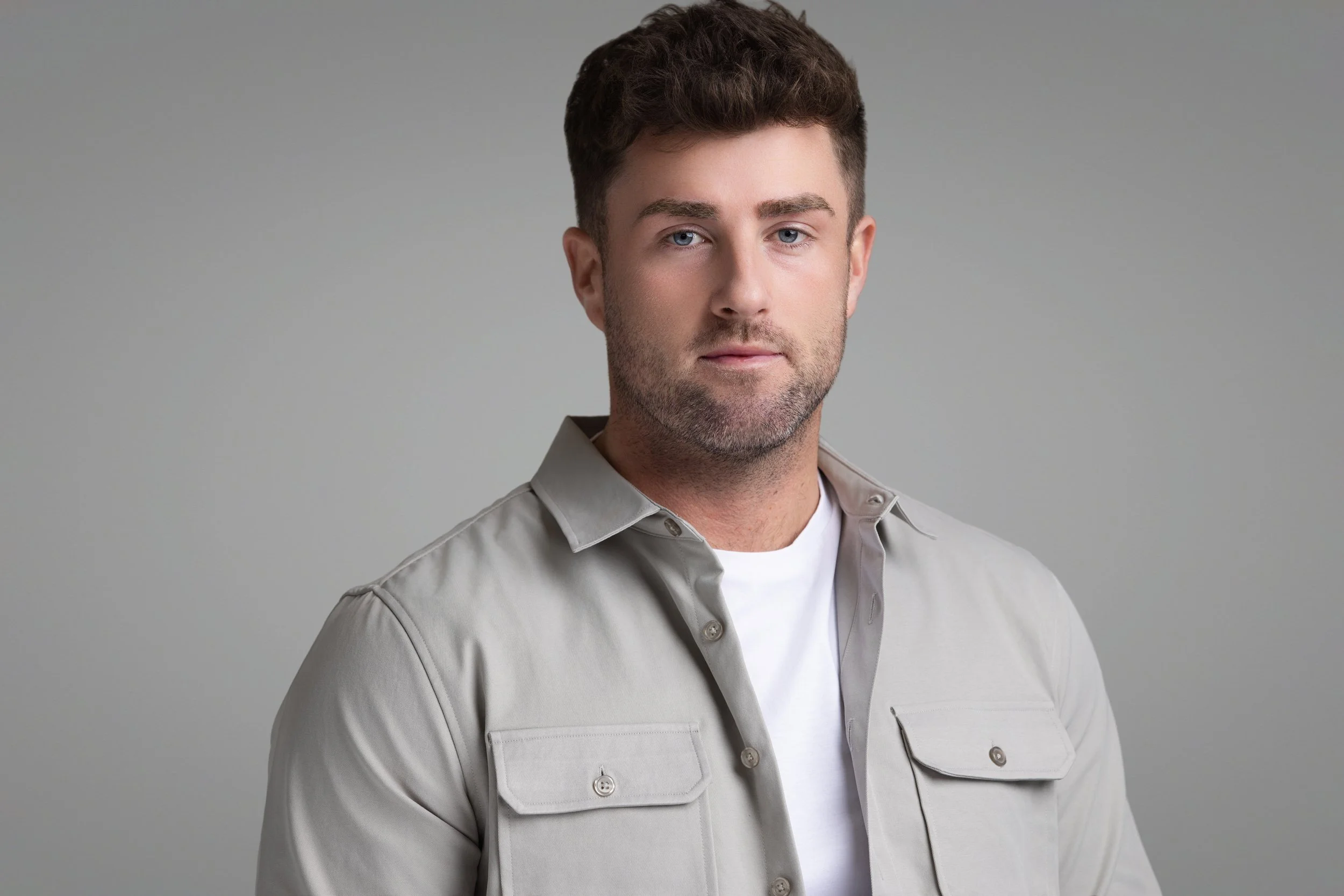 A man with short dark hair and blue eyes, wearing a light-colored button-up shirt over a white T-shirt, looking directly at the camera against a plain gray background.