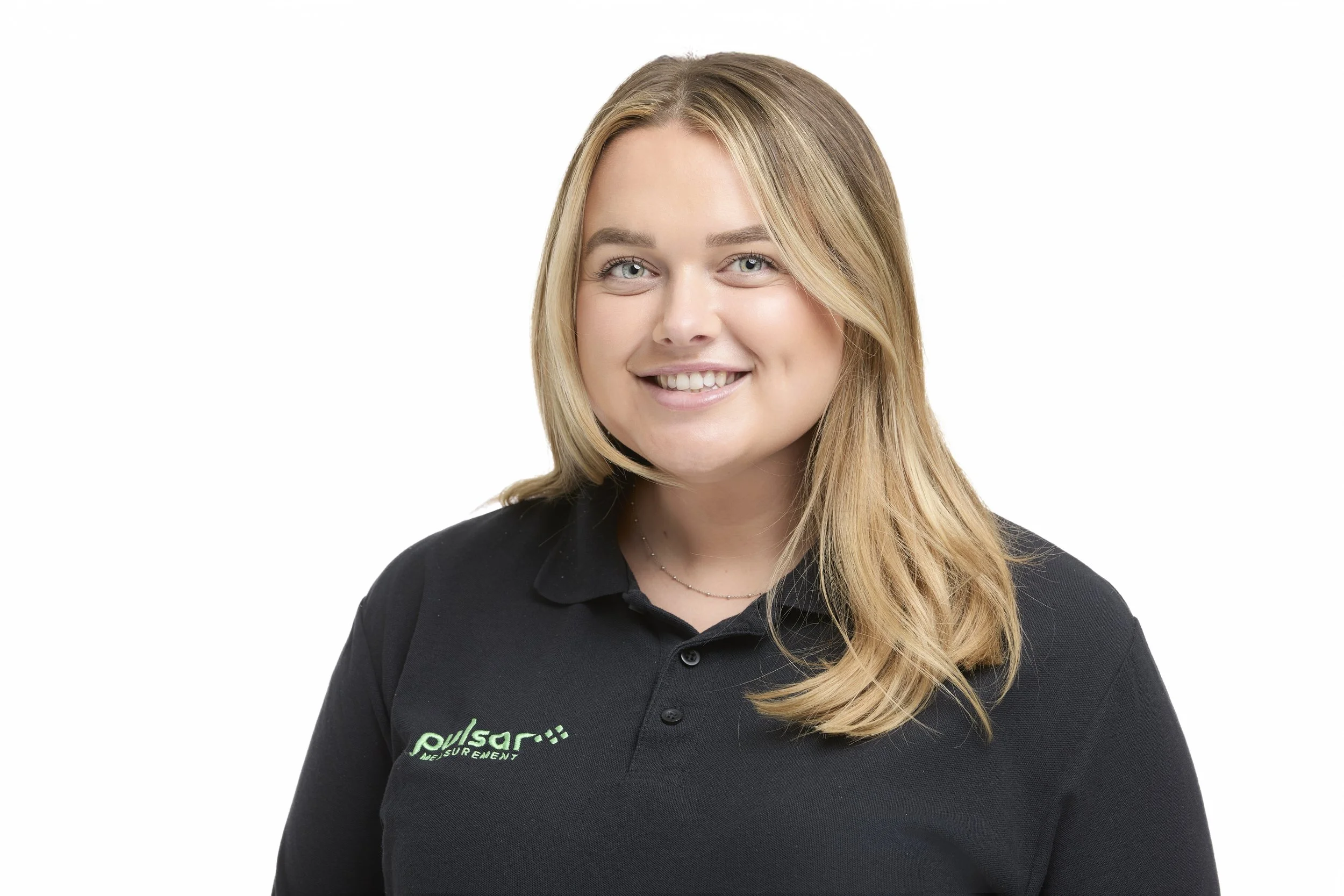 A young woman with blonde hair smiling, wearing a black collared shirt with green embroidered logo, against a white background.
