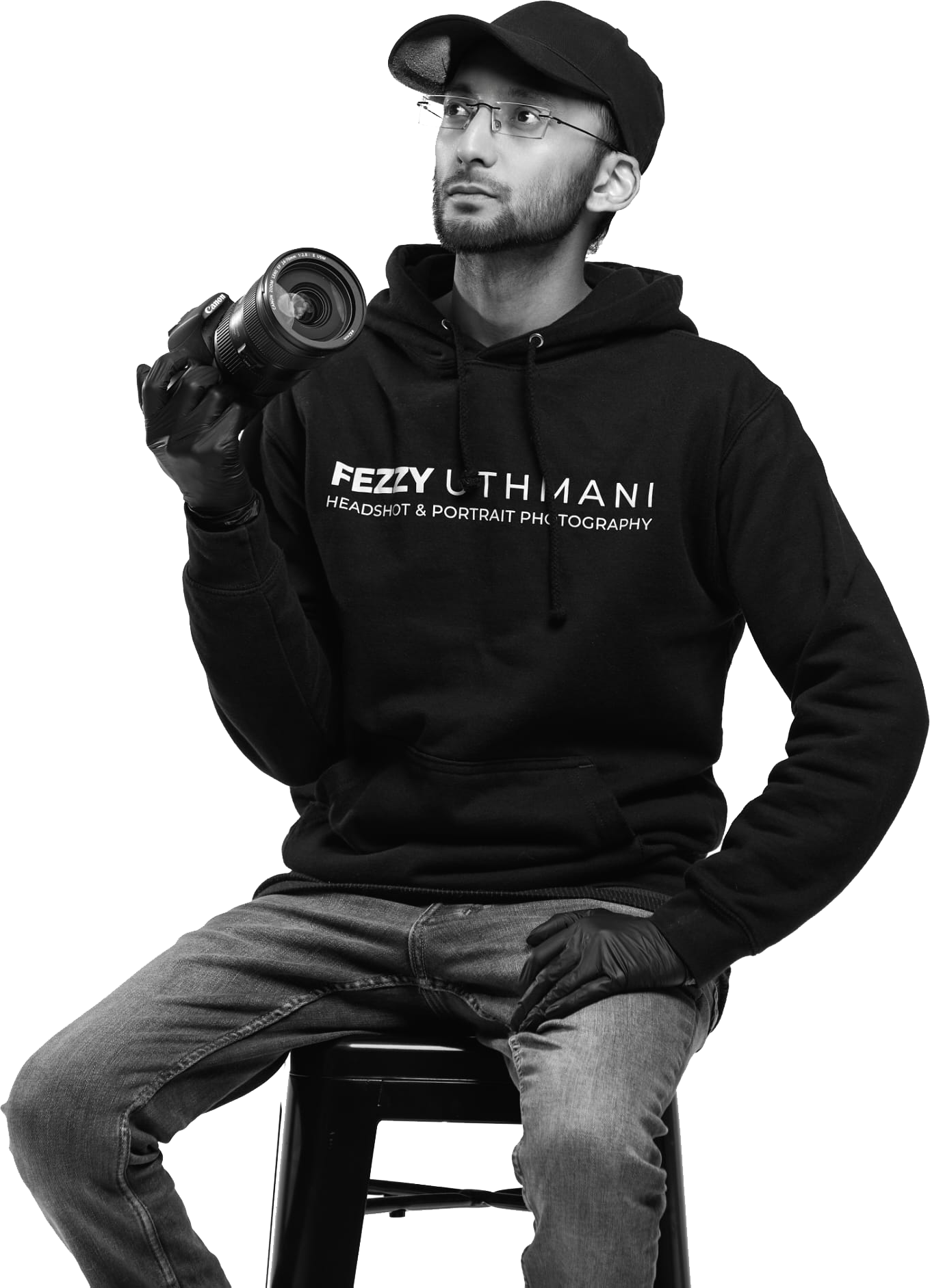 A man wearing a black hoodie, glasses, and a cap, sitting on a stool, holding a camera lens in his right hand, and wearing a glove on that hand, with a serious expression.