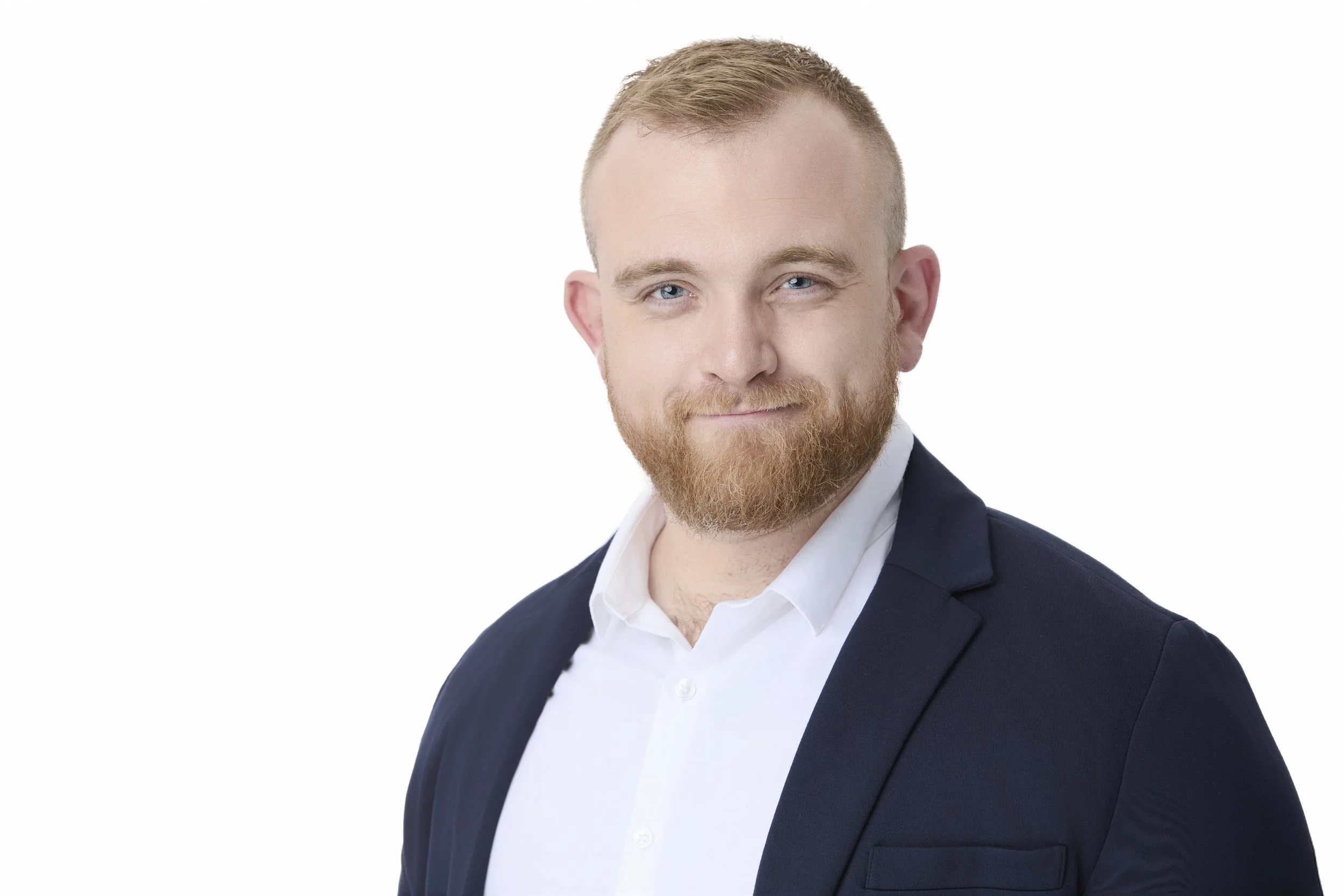 Headshot of a young man with blonde hair, a beard, wearing a navy blazer and white shirt, smiling slightly against a white background.