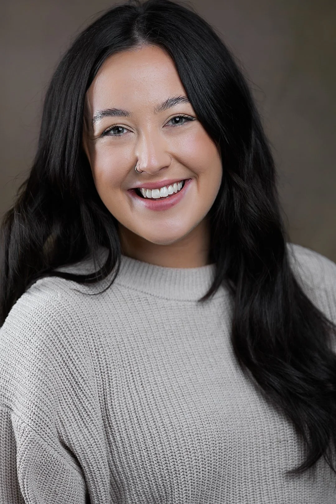 A woman with long dark hair, smiling, wearing a beige knit sweater, against a neutral background.