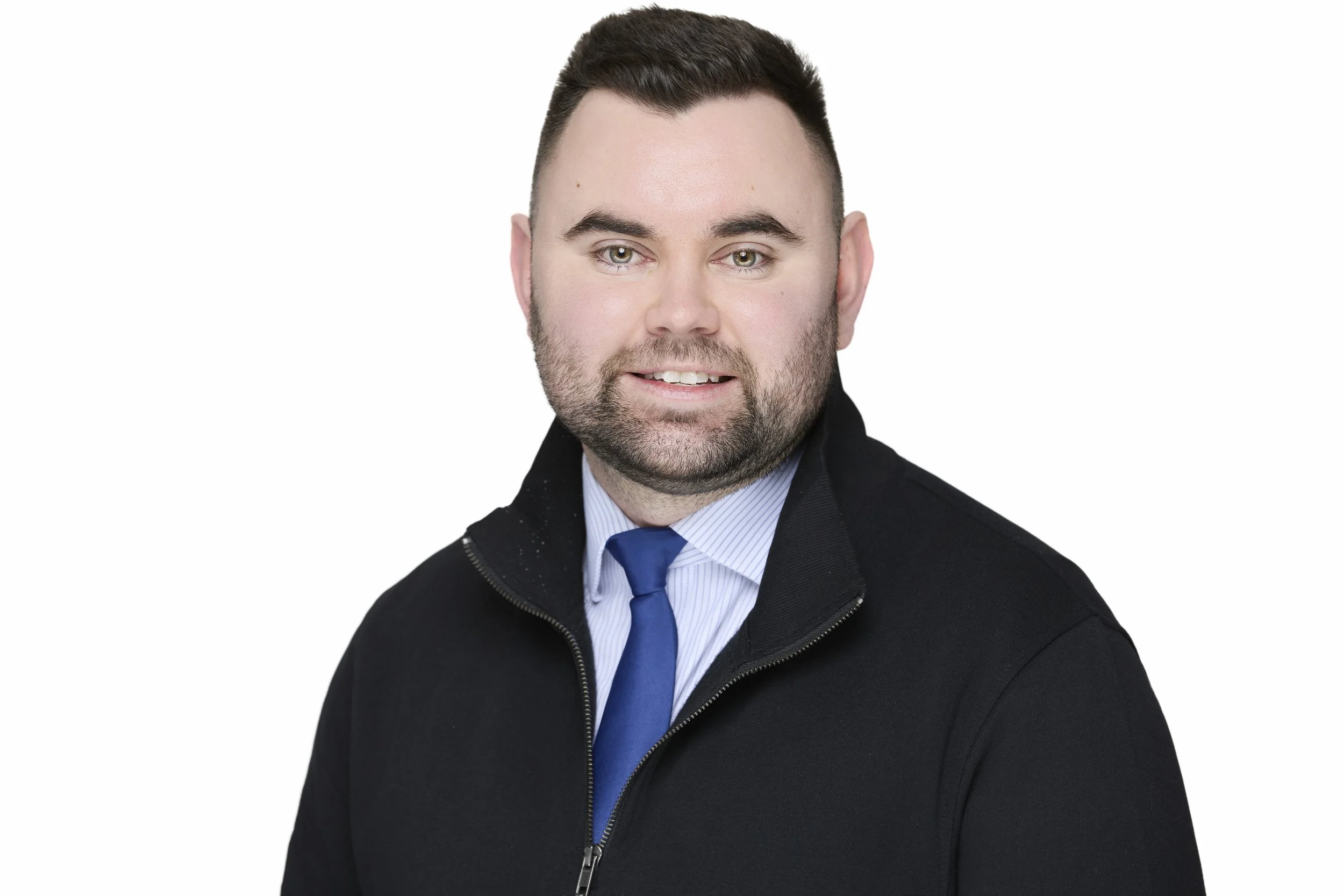 Professional headshot of a man with short dark hair and a beard, wearing a black jacket over a blue shirt and a blue tie, against a plain white background.