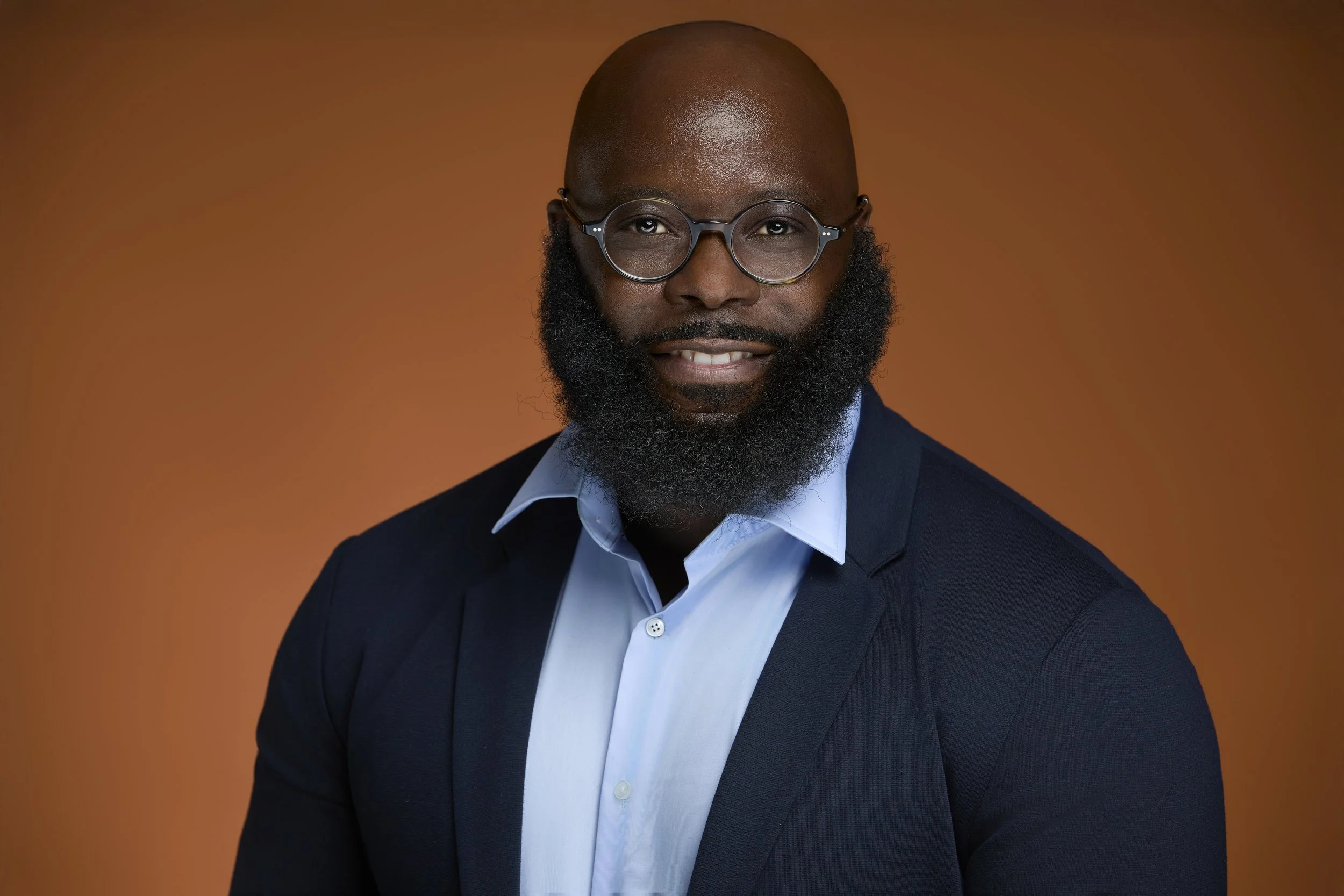 A man wearing glasses in a dark blazer and a white shirt poses against a warm orange background.