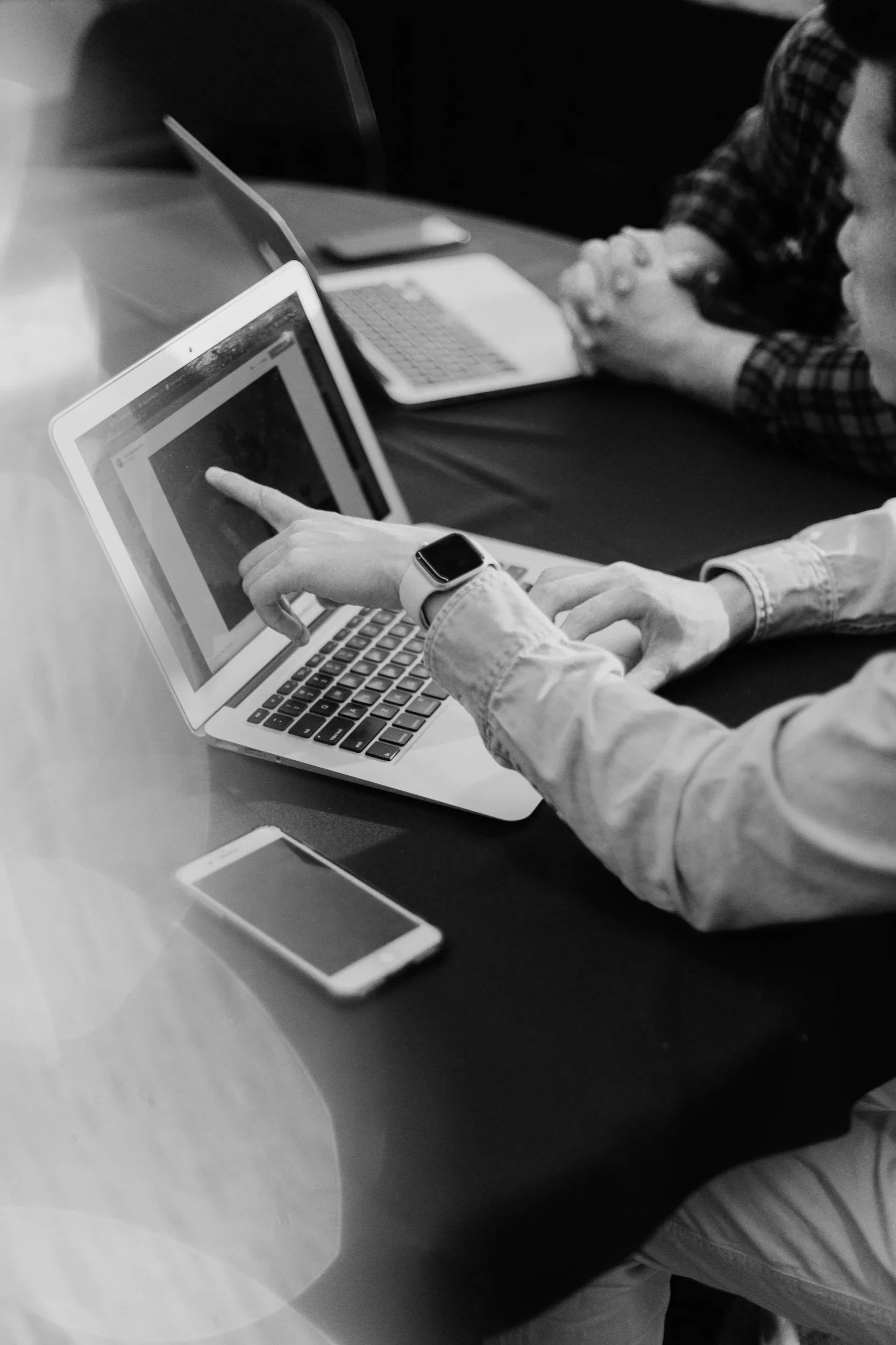 A person using a laptop, pointing at the screen, with a smartphone on the table nearby. Other individuals are seated at the table, engaged in work or discussion.