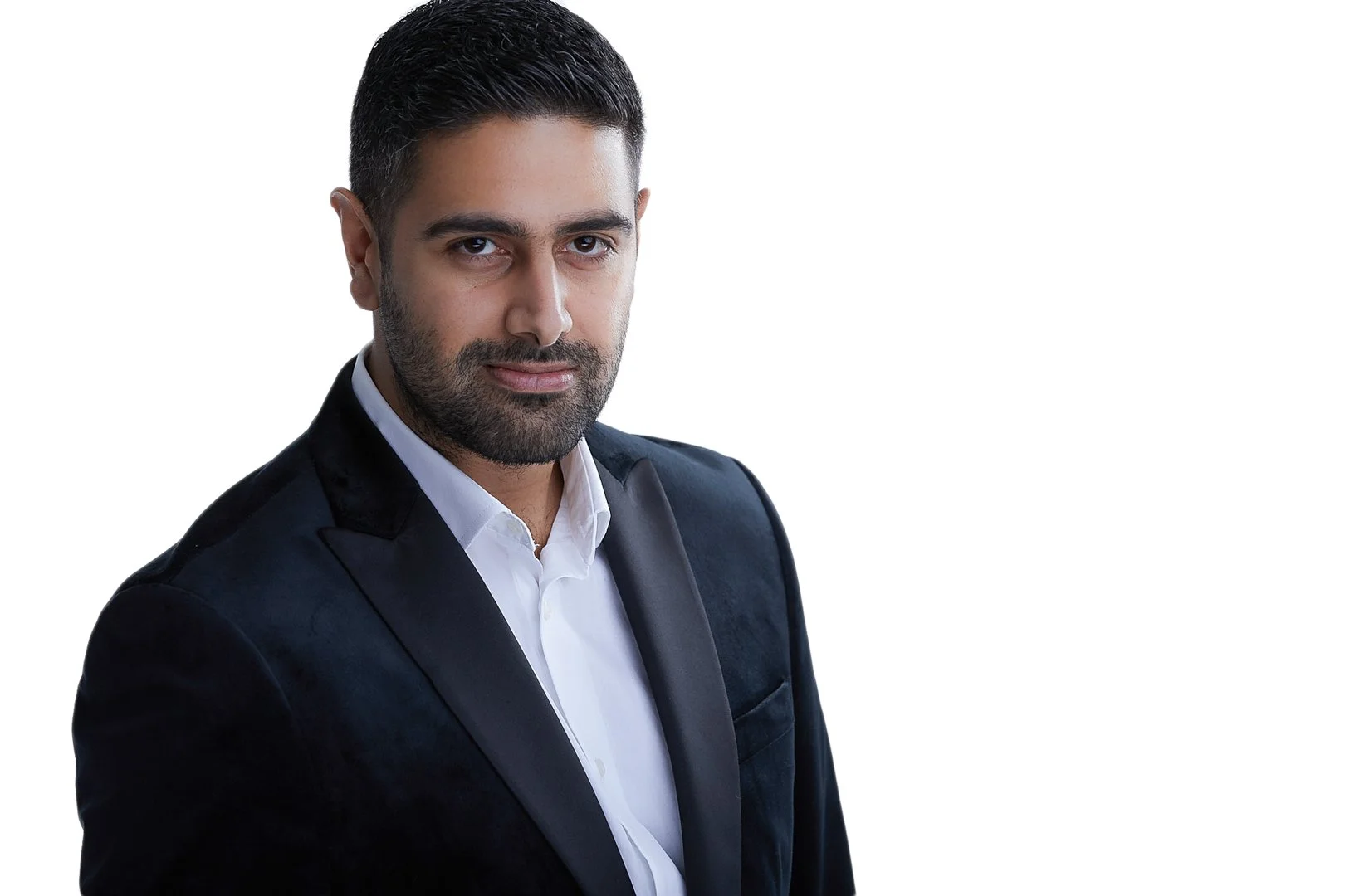 A man with short dark hair and a beard, wearing a black suit and white dress shirt, looking at the camera against a plain white background.
