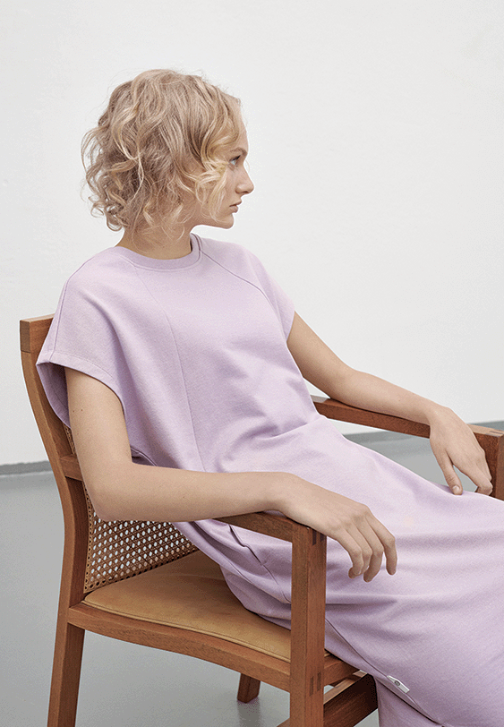 A young woman with curly blonde hair sits sideways on a wooden chair with a cane backrest, wearing a light purple dress, in a minimalistic room with white walls.